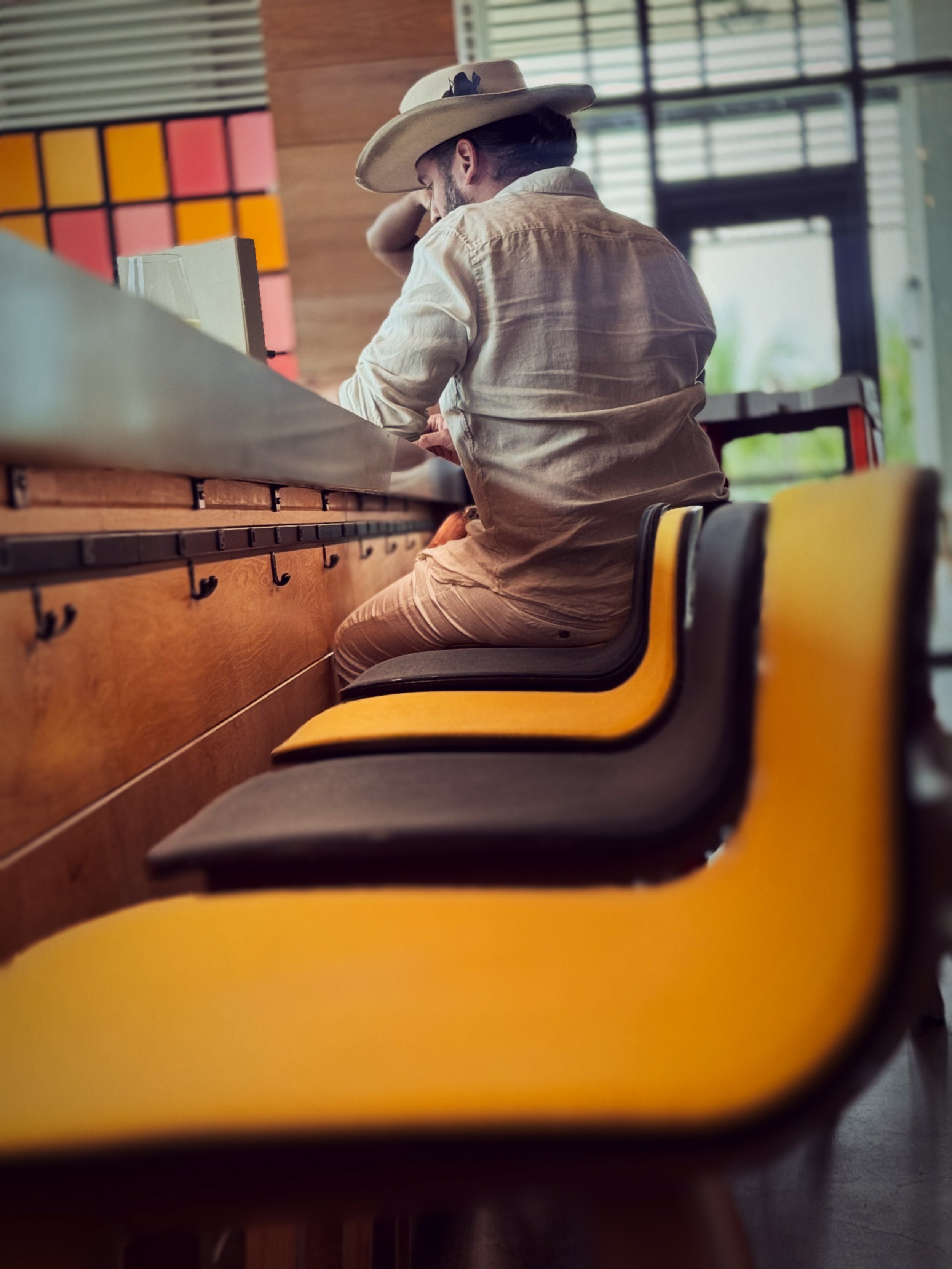 A man wearing a wide-brimmed hat and beige clothing is sitting on a yellow and black chair at a wooden counter inside a cafe or restaurant, with books and colorful square wall art behind him.
