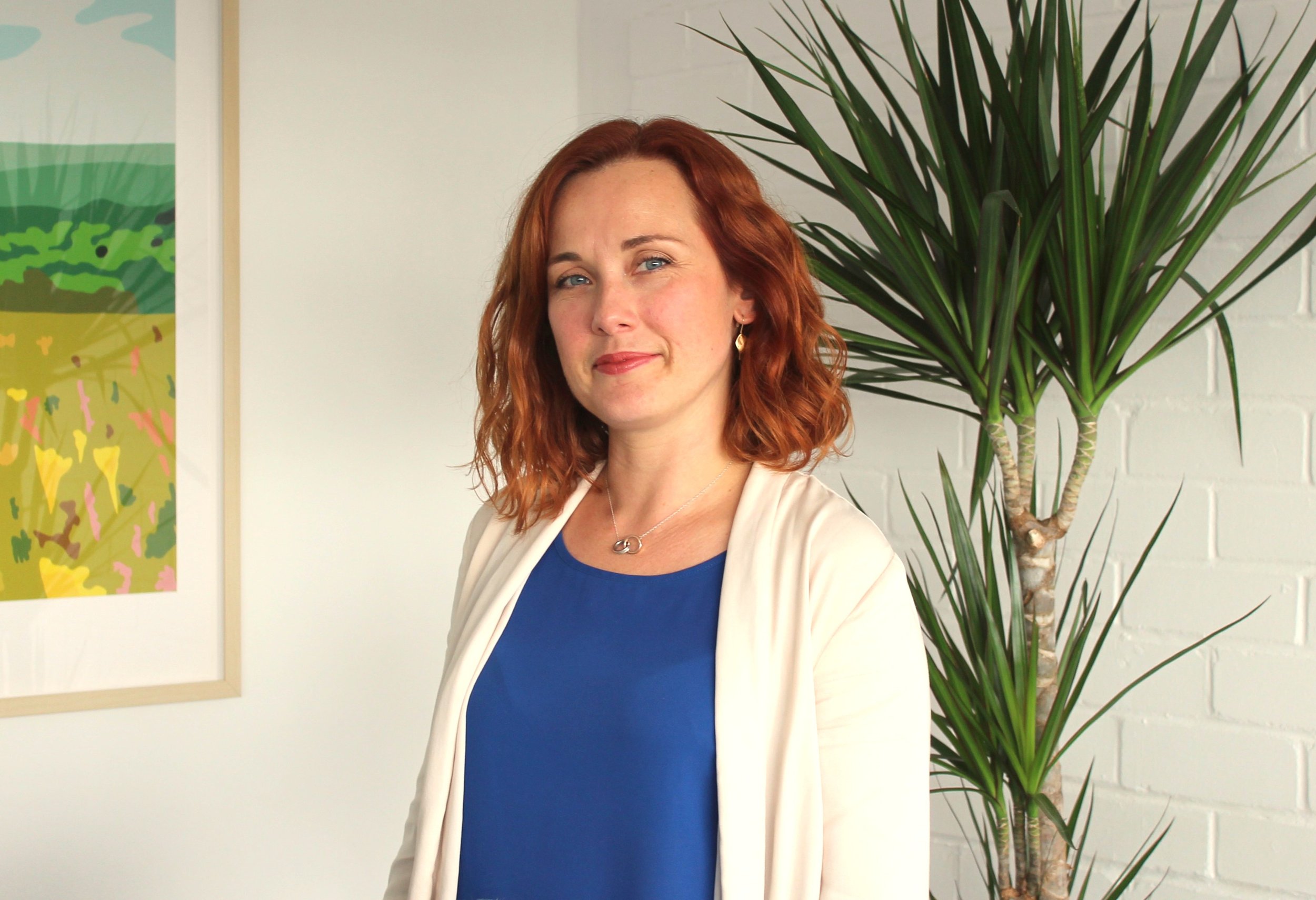 A woman with red hair, light skin, and blue eyes standing indoors next to a large green plant, white brick wall, and colorful abstract art on the wall.