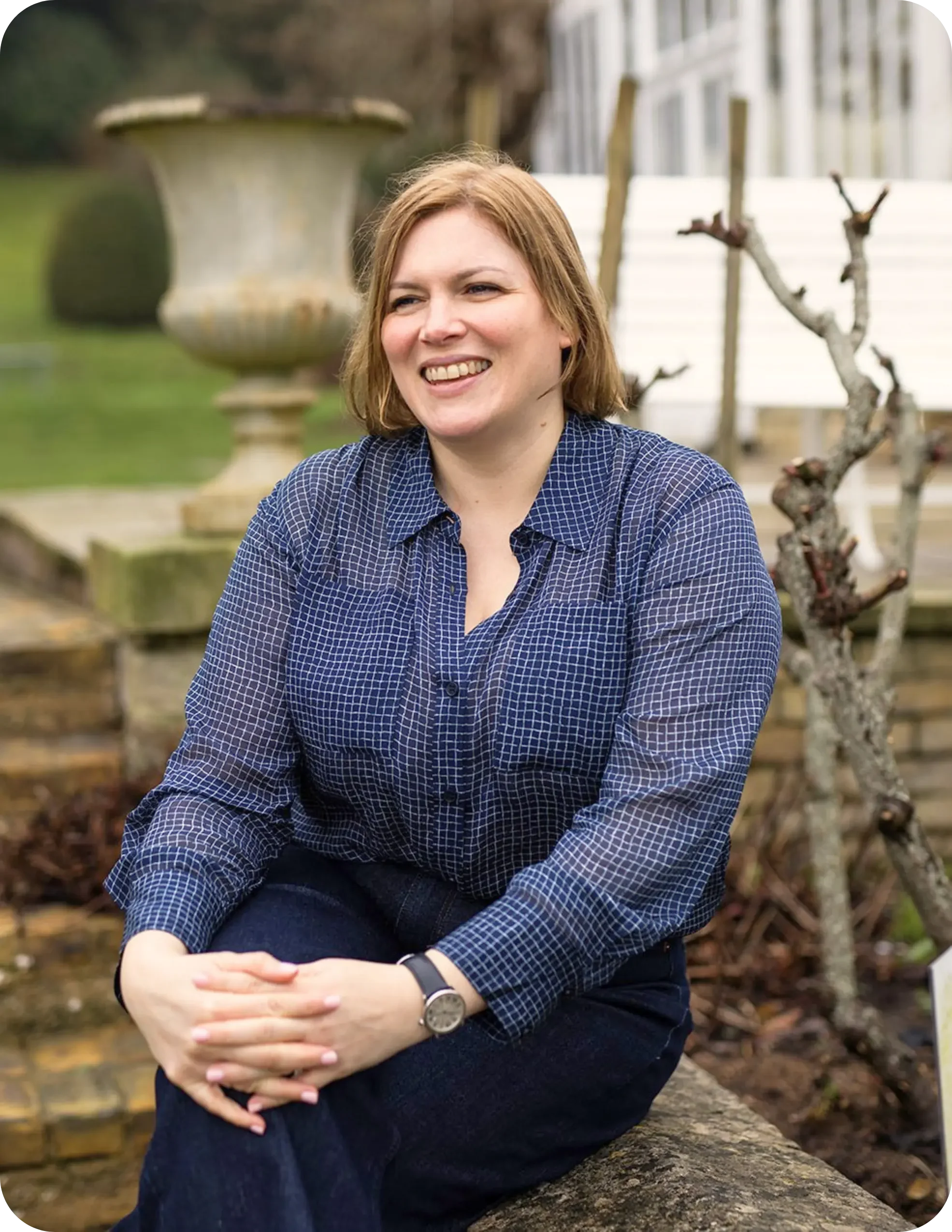 A woman with short brown hair and light skin sitting outdoors, smiling, wearing a blue checkered shirt and a silver watch, with a plant pot and a tree in the background.
