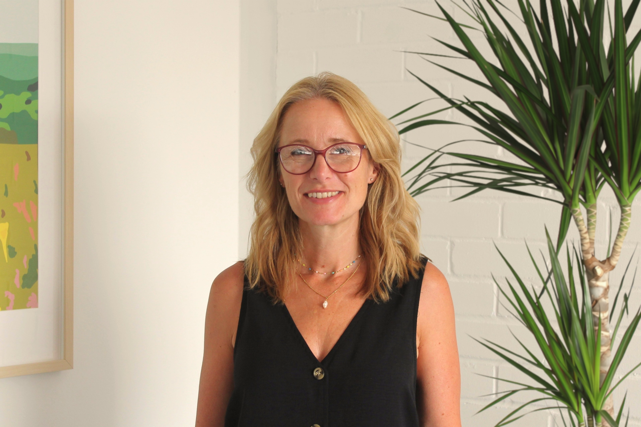 A woman with blonde hair wearing glasses and a sleeveless black top, smiling, standing indoors next to a large green plant, with artwork on the wall behind her.