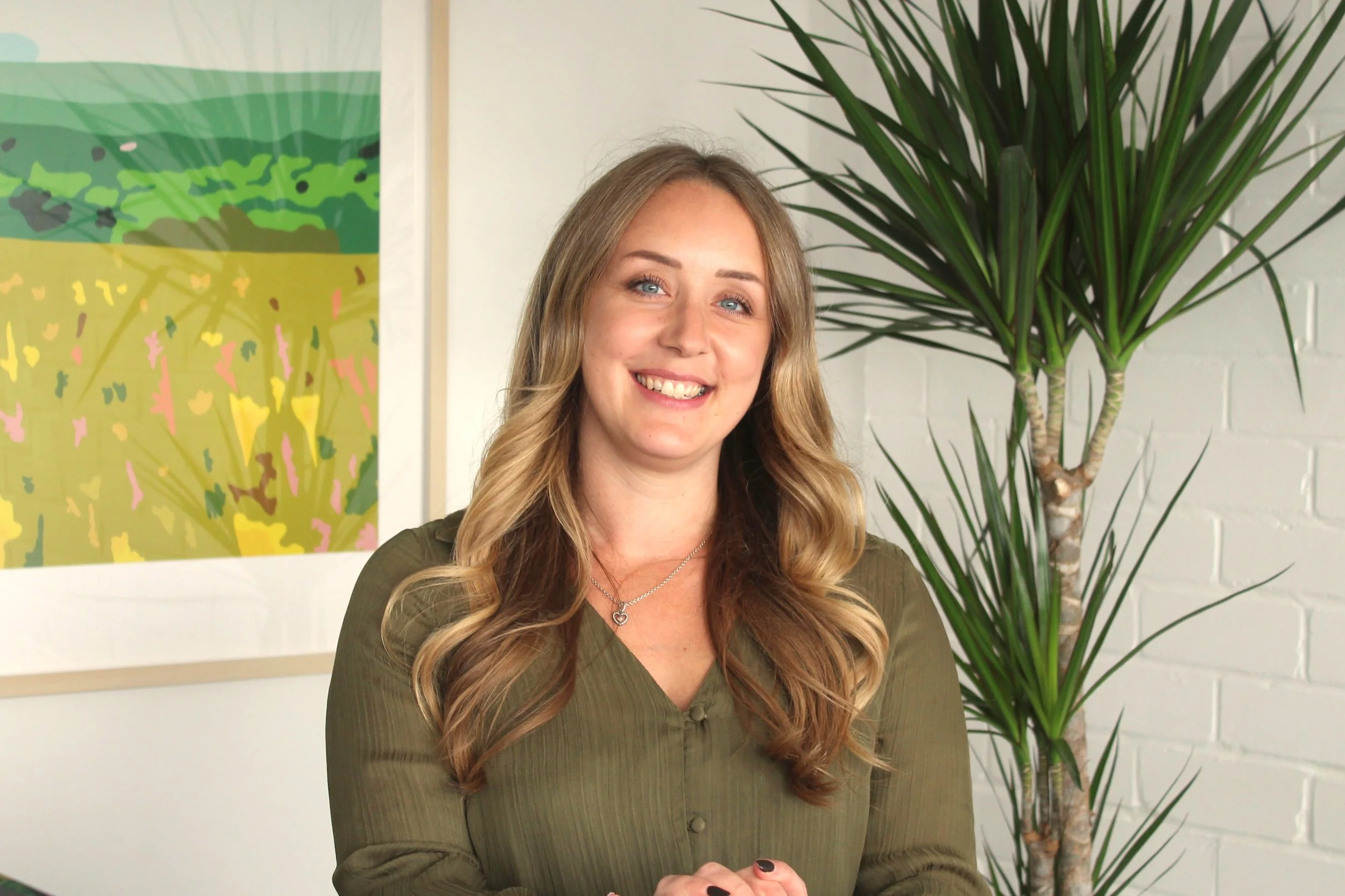 A smiling woman with long wavy blonde hair, wearing a green top and a heart-shaped necklace, standing in front of a large indoor plant and a colorful abstract painting on a white wall.