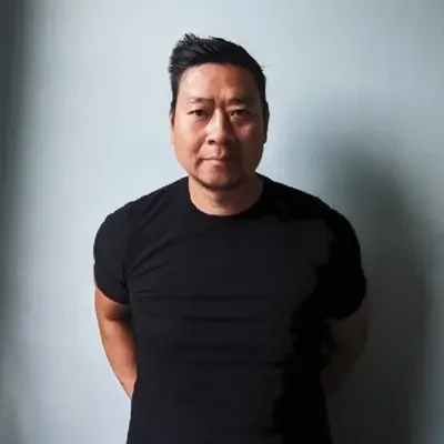 A man with short black hair wearing a black T-shirt, standing against a plain light-colored wall.
