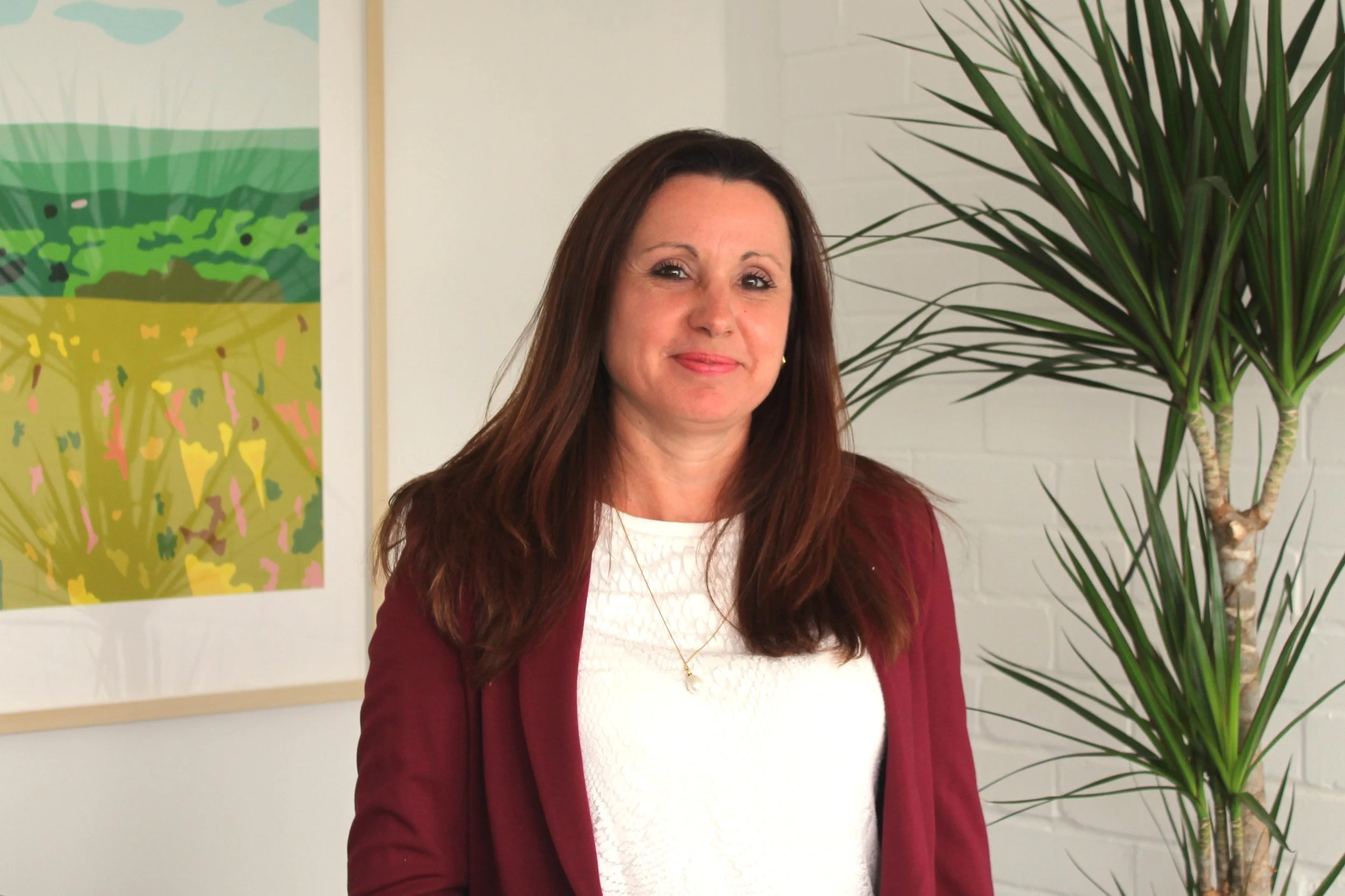 A woman with long brown hair, wearing a white top and a maroon blazer, is standing indoors near a large potted plant with long green leaves. There is a colorful, abstract painting on the wall behind her.