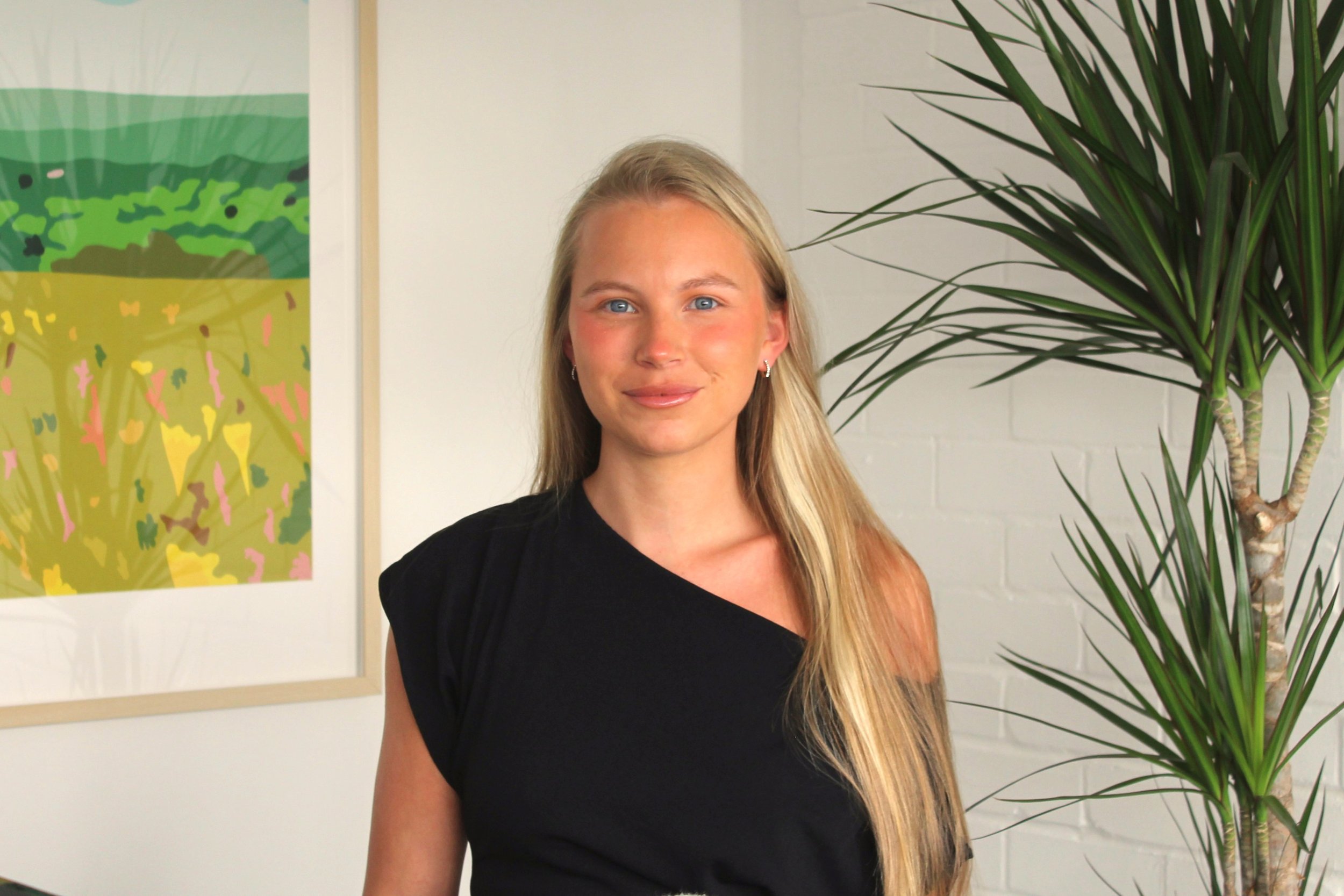 Young woman with long blonde hair and blue eyes standing indoors in front of a colorful abstract artwork and a tall green plant, smiling at the camera.