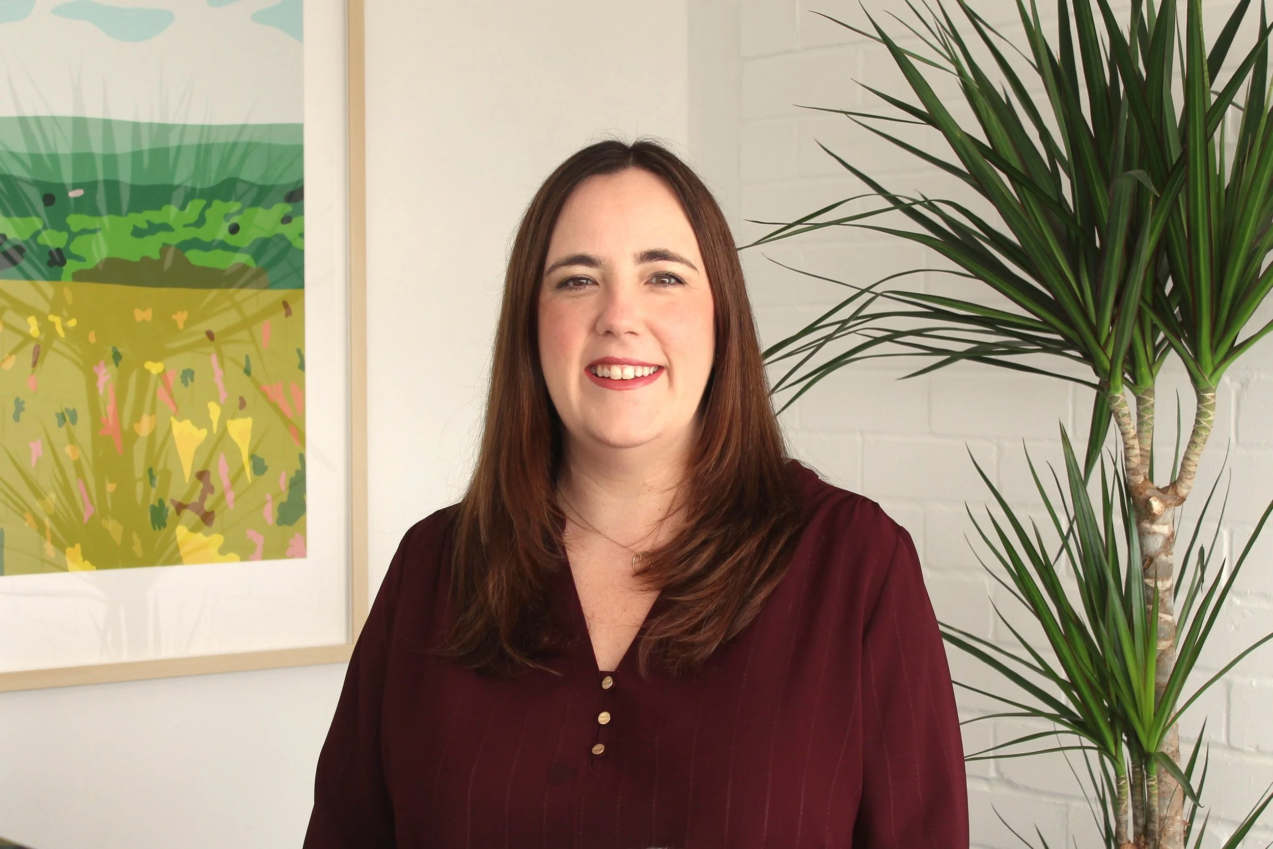 A woman with long brown hair smiling, wearing a maroon blouse, standing indoors near a large green potted plant and colorful abstract artwork on the wall.