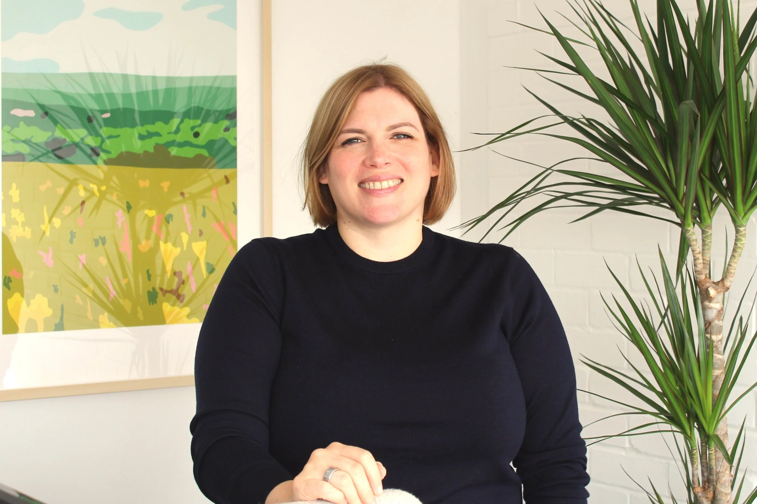 A woman with short red hair and a black sweater is smiling, sitting indoors next to a tall green plant with spiky leaves. There is colorful abstract art on the wall behind her.