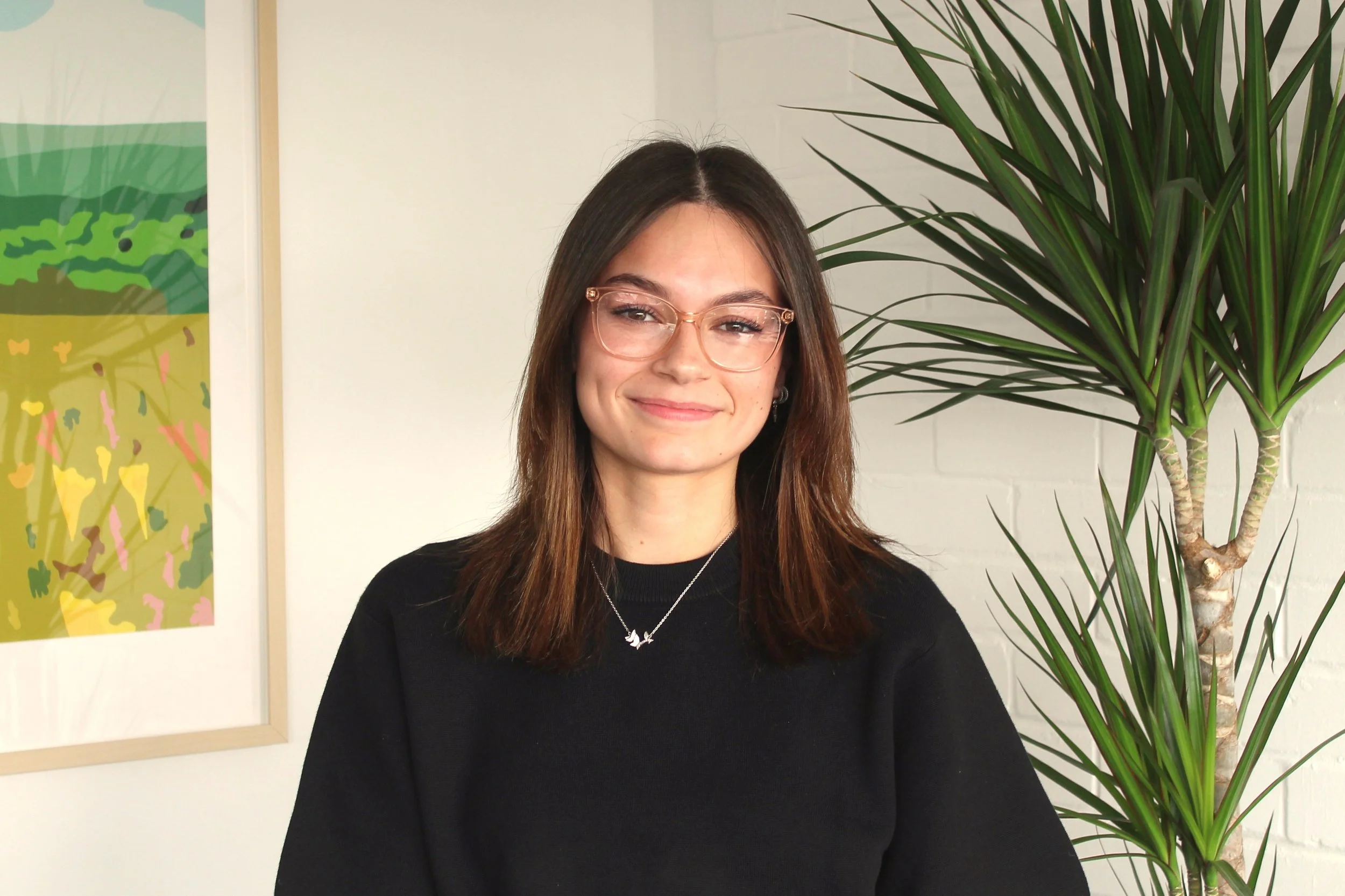 A woman with long brown hair wearing glasses and a black top, standing indoors next to a large green plant, with colorful artwork on the wall behind her.