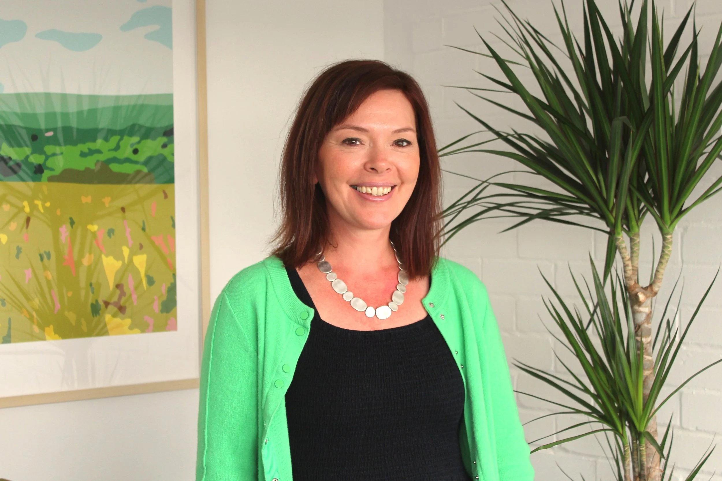 A woman with shoulder-length brown hair smiling, wearing a bright green cardigan, black top, and a white circular necklace, standing next to a large green plant with long, slender leaves, in front of a white wall and colorful abstract artwork.