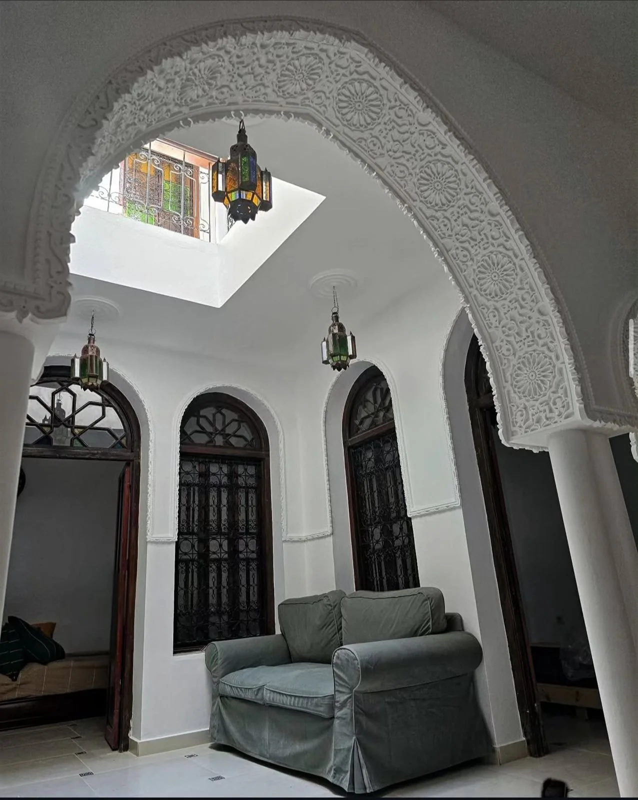 Interior of a room with white walls, arched windows with black wrought iron grilles, and a gray velvet sofa. The ceiling features a skylight and decorative hanging lanterns, with intricate white plasterwork around the arches.
