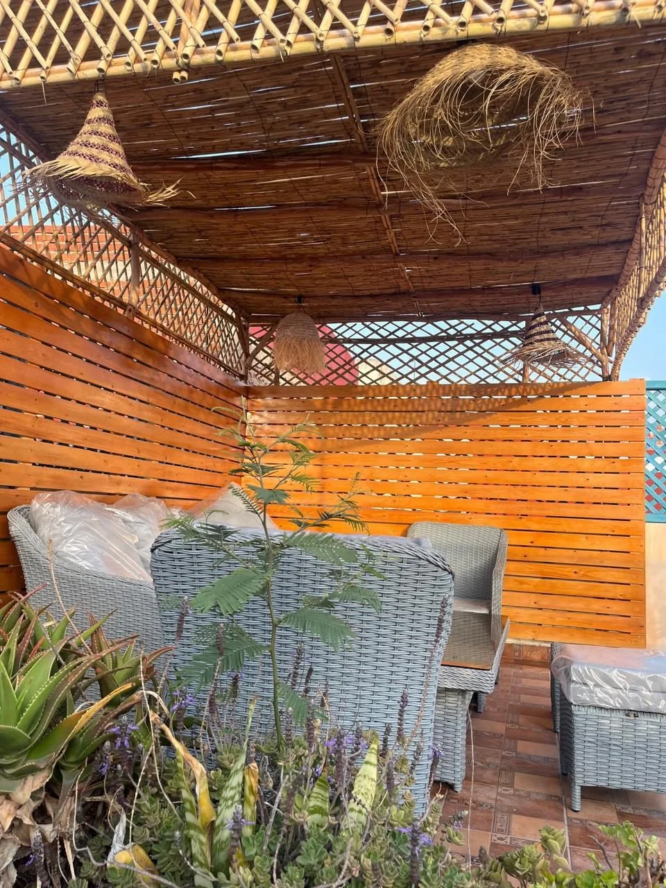 Outdoor shaded patio space with wicker furniture, potted plants, and woven hanging decorations under a bamboo roof.