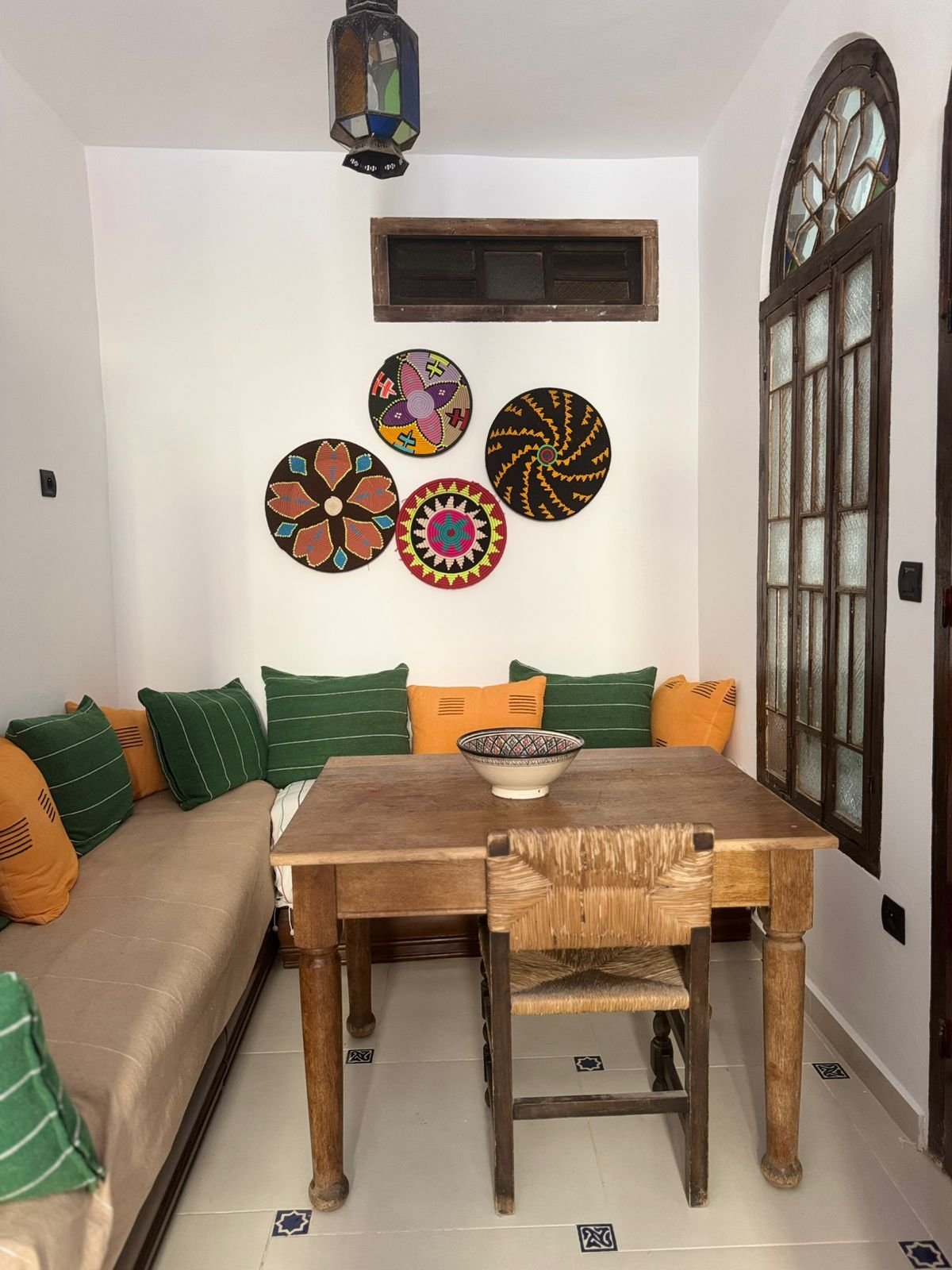 A cozy dining corner with a wooden table, surrounded by green and orange cushions, on a white tiled floor with small decorative tile accents. There are colorful woven wall hangings above the table, and a stained glass window with a wooden frame on th