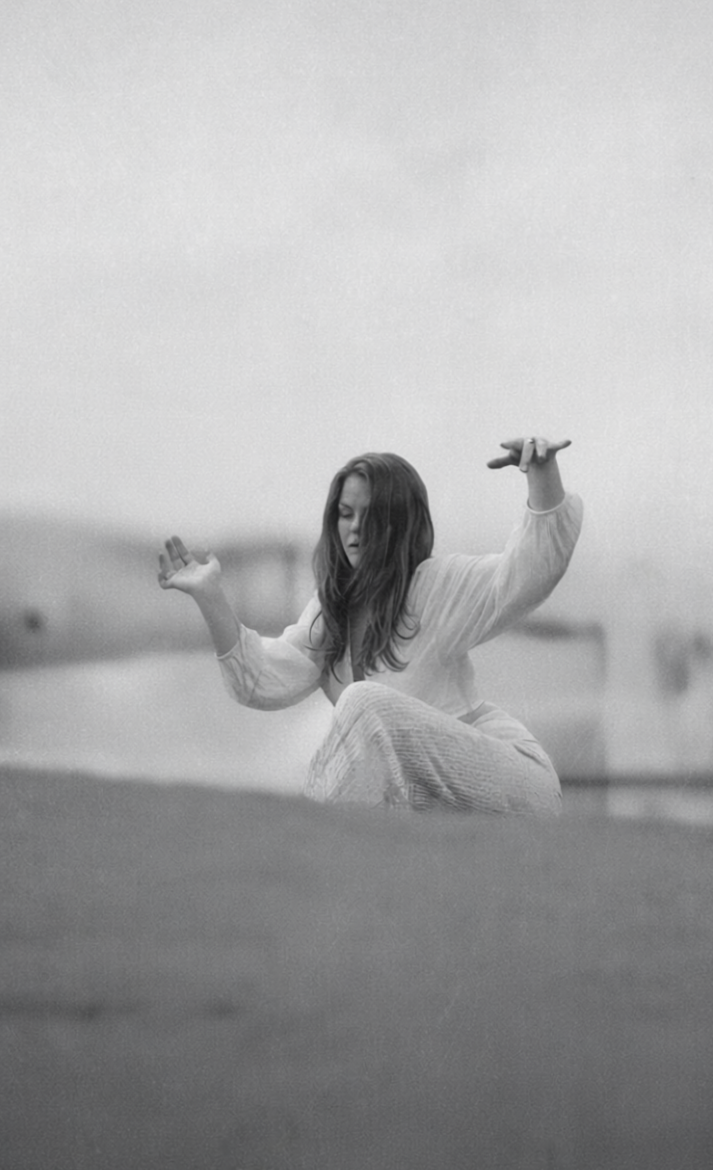 Schwarz-weiß-Foto einer Frau, die in einer meditativen Haltung sitzt, die Augen geschlossen hat und die Hände in einer entspannten Position hält, auf einer flachen Oberfläche im Freien.