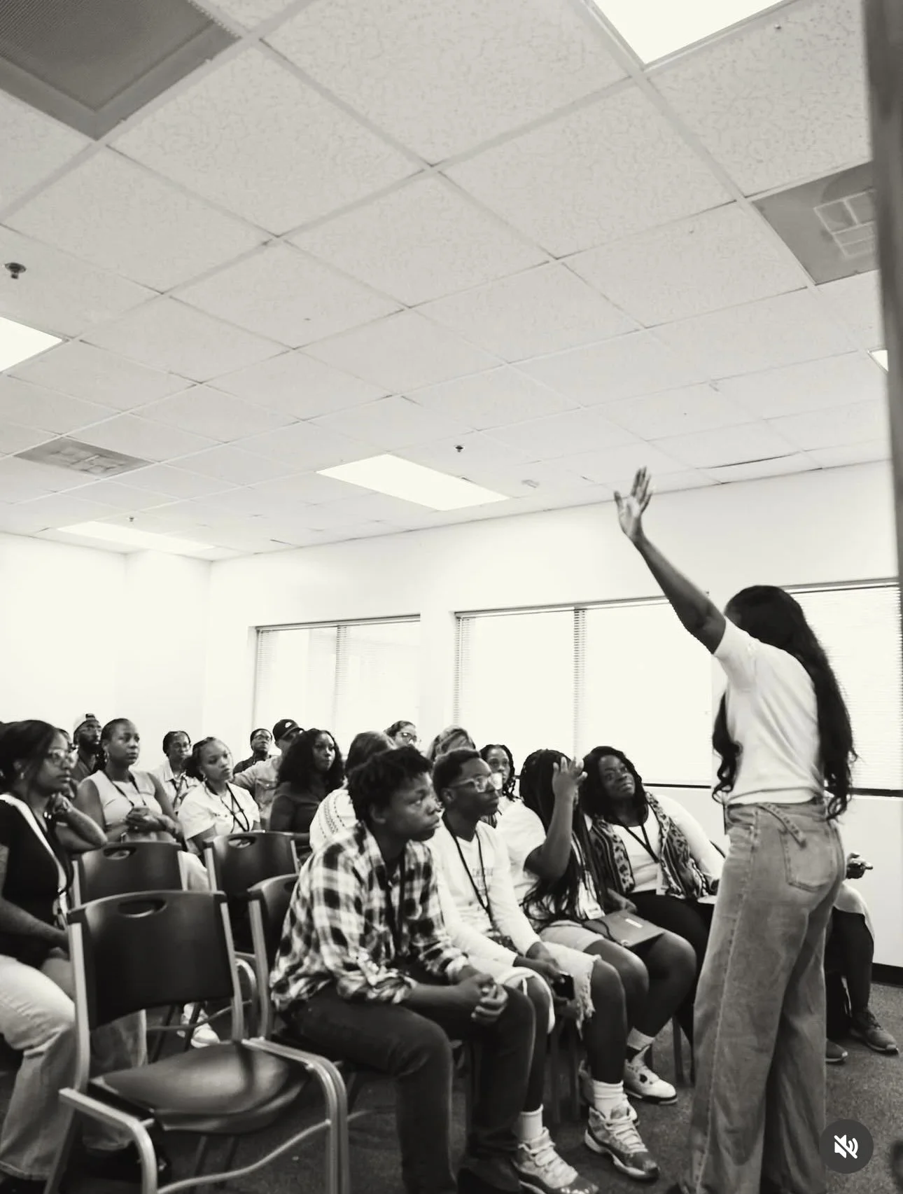 A woman is leading a discussion or presentation in front of a group of young women and girls in a classroom or conference room.