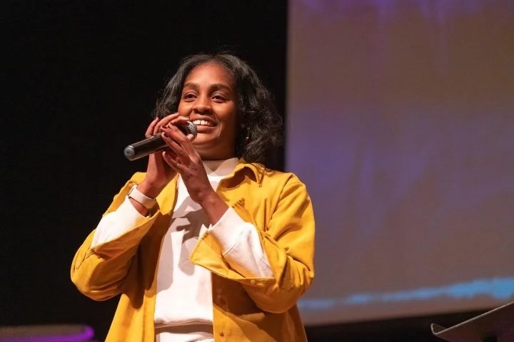 A young woman with shoulder-length curly hair, wearing a mustard yellow jacket over a white shirt, smiling and holding a microphone on stage.