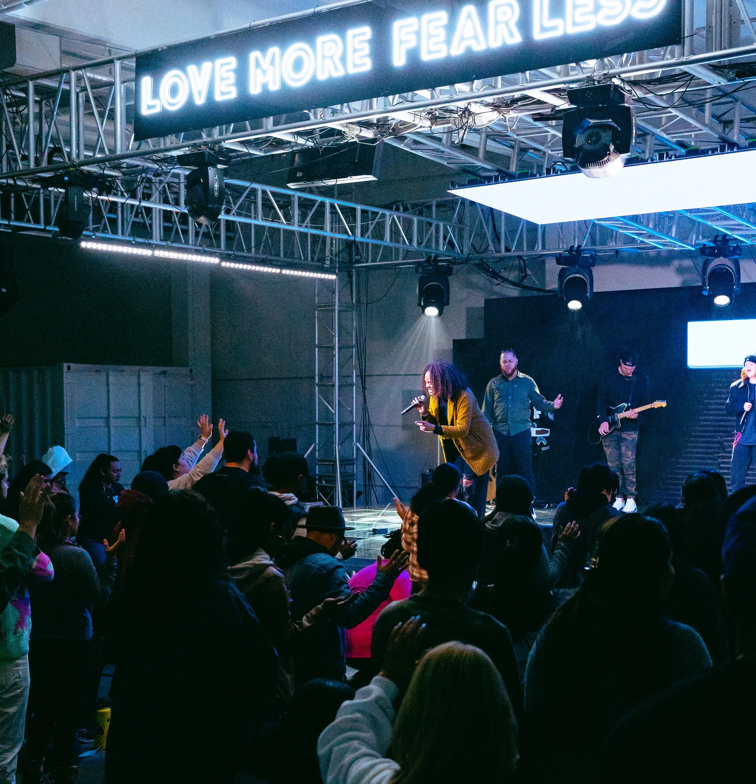 A woman speaking in a microphone on a stage with band members, audience raising hands, neon sign reads 'Love More Fear Less'.