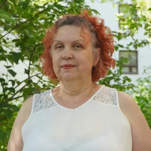 Nainen hymyilee ja pitää kahvikuppia ja kansiota, seisoo sisätila-alueella, taustalla kasveja ja ikkunat.