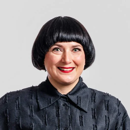 Nainen, jolla lyhyt musta bob-letti ja punahuulikasit, hymyilee studiossa harmaata taustaa vasten.