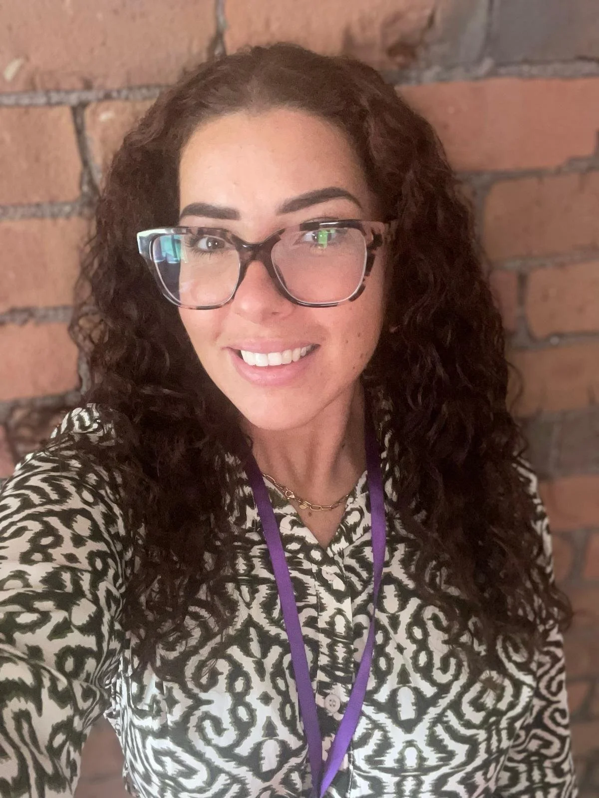 A woman with curly brown hair and glasses taking a selfie against a brick wall, wearing a patterned black and white top and a purple lanyard.