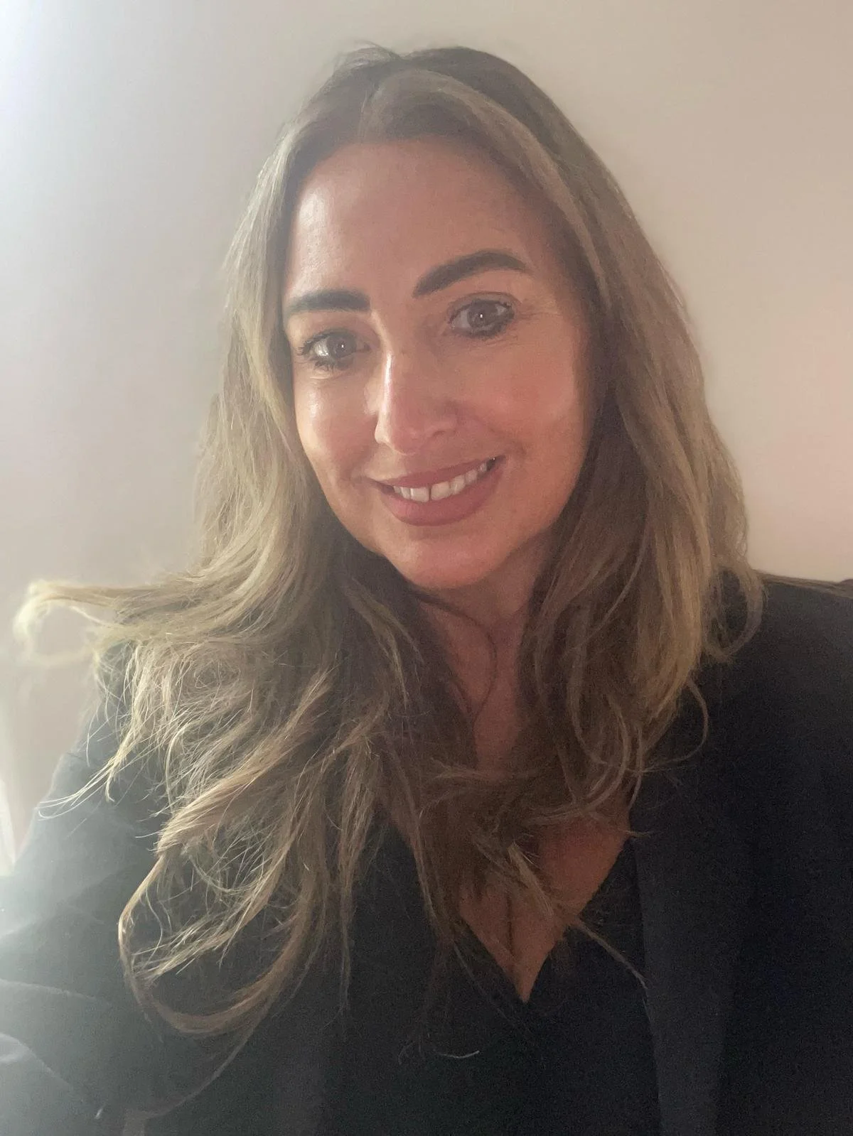 A woman with long, wavy brown hair smiling in front of a plain wall.