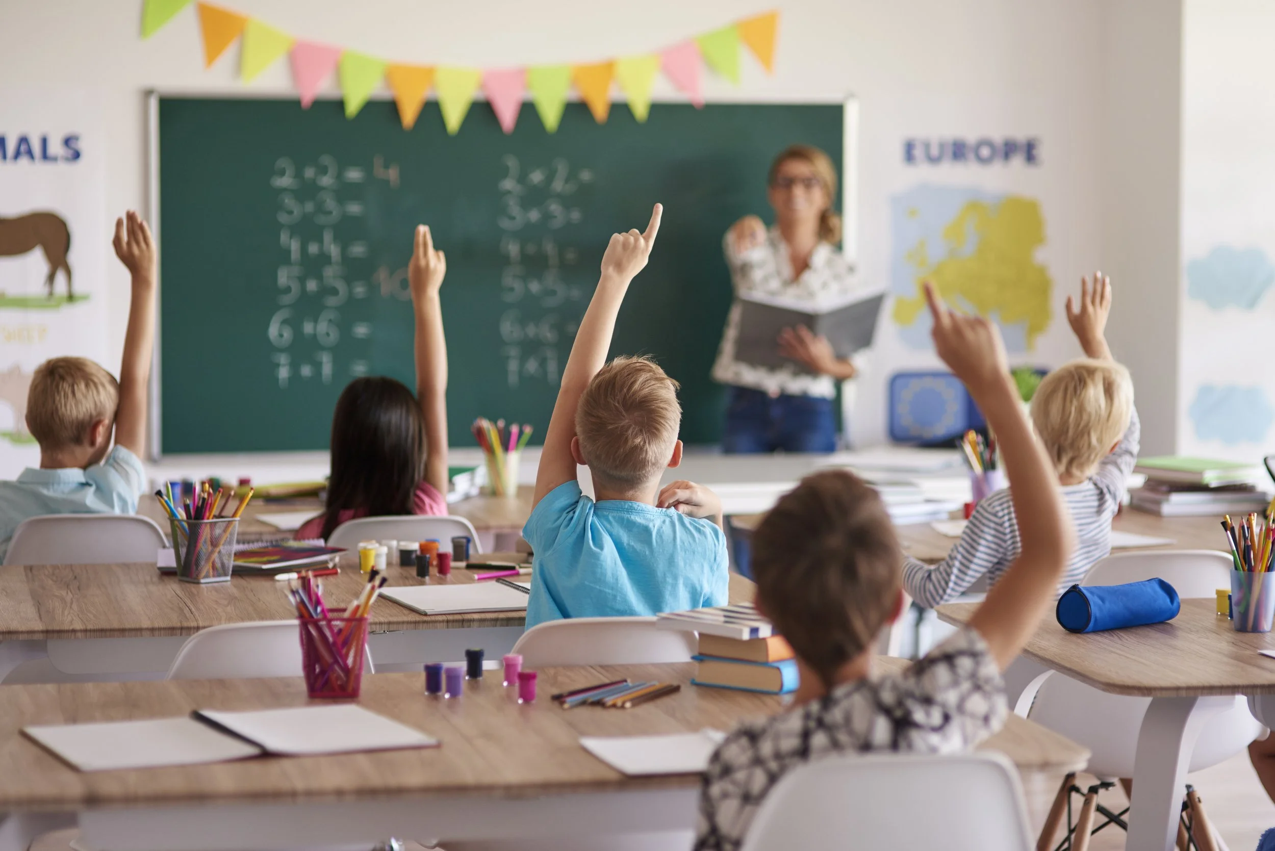 Classroom with young students raising hands at their desks, teacher at blackboard with math exercises, colorful decorations, and educational posters.