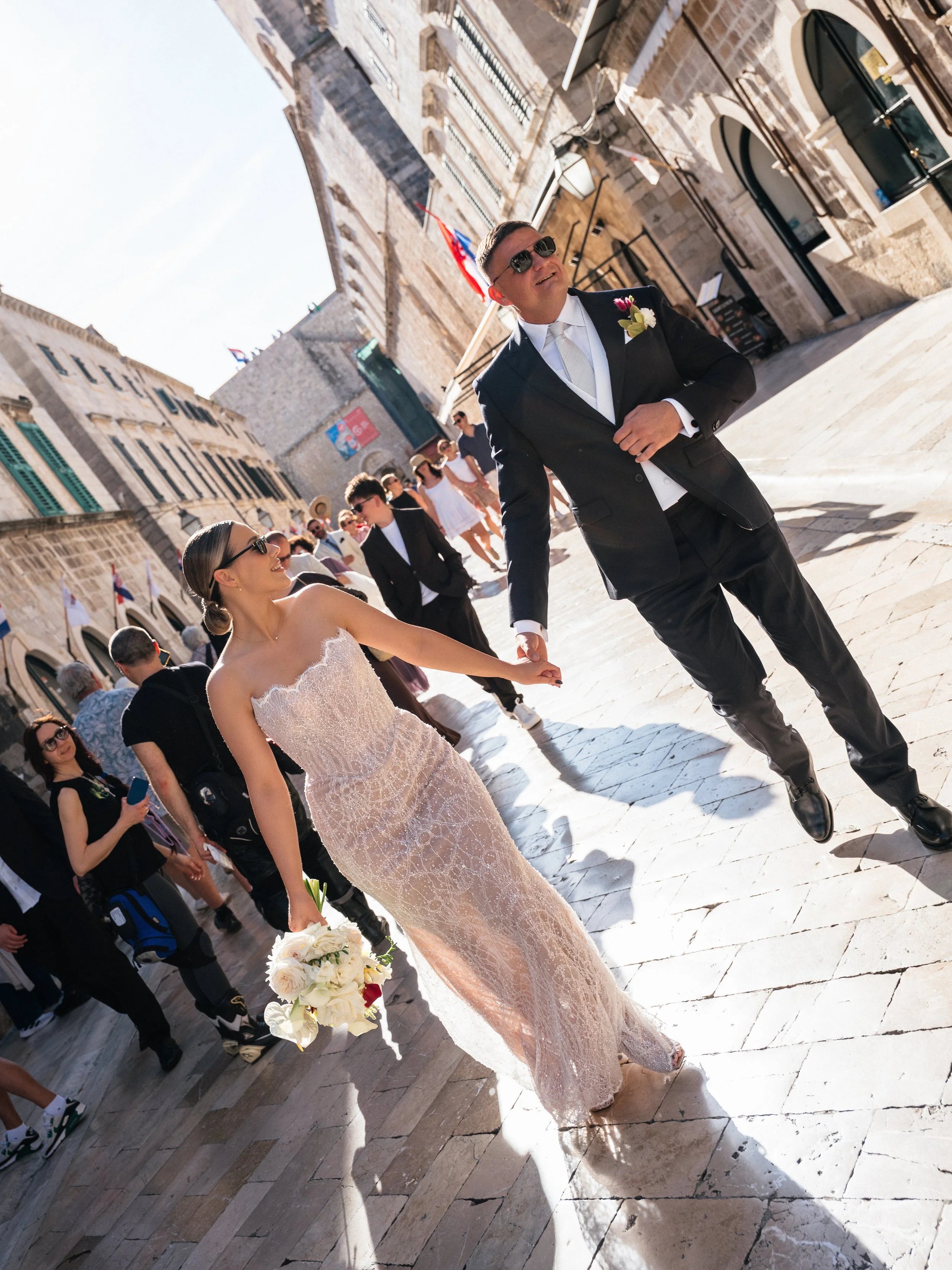 A man and woman holding hands and walking on a cobblestone street, likely during a wedding or special event, with other people in the background and historic buildings lining the street.