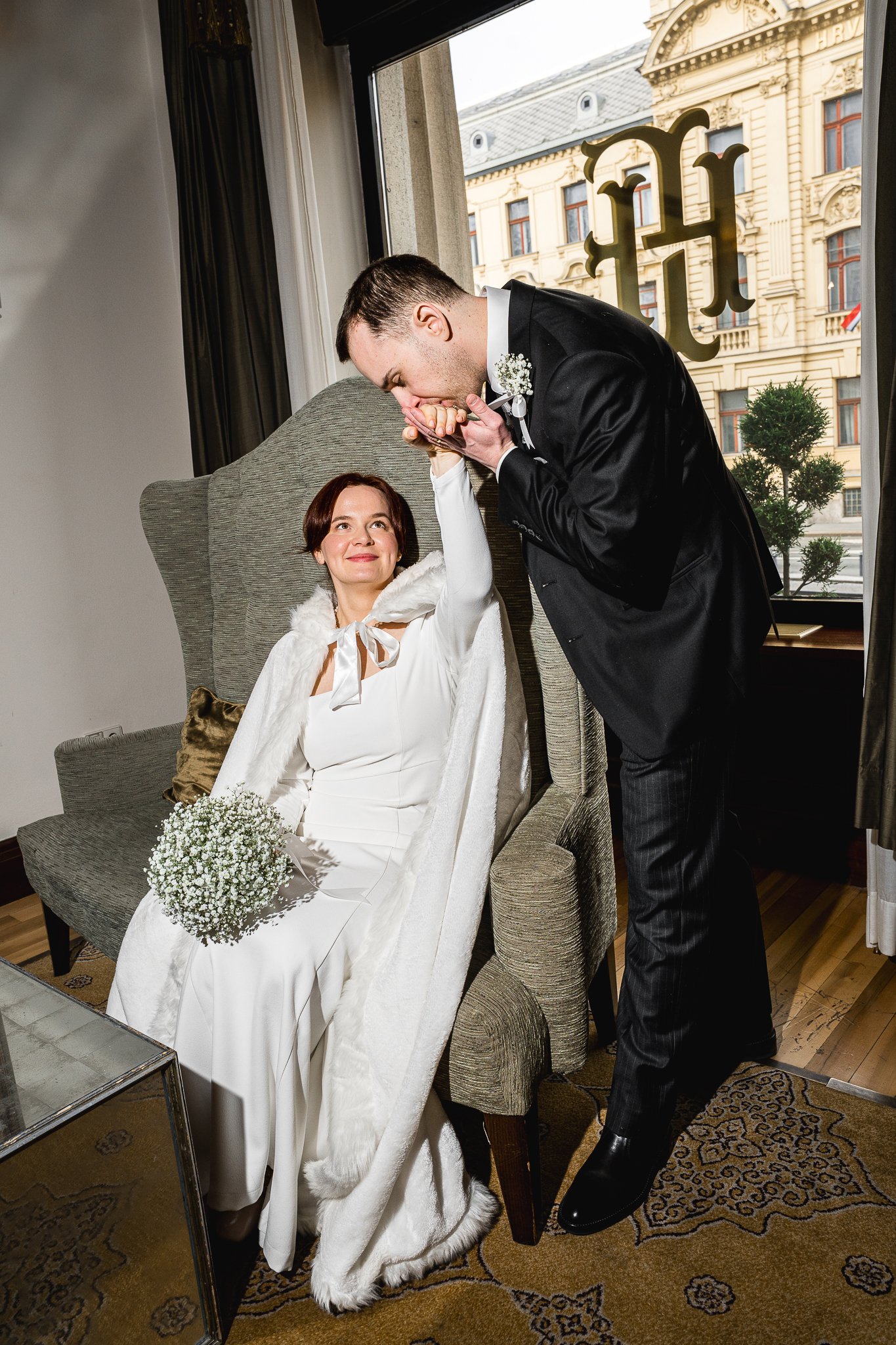 A groom kissing a bride on the hand in an indoor setting with a large window showing an ornate building outside.