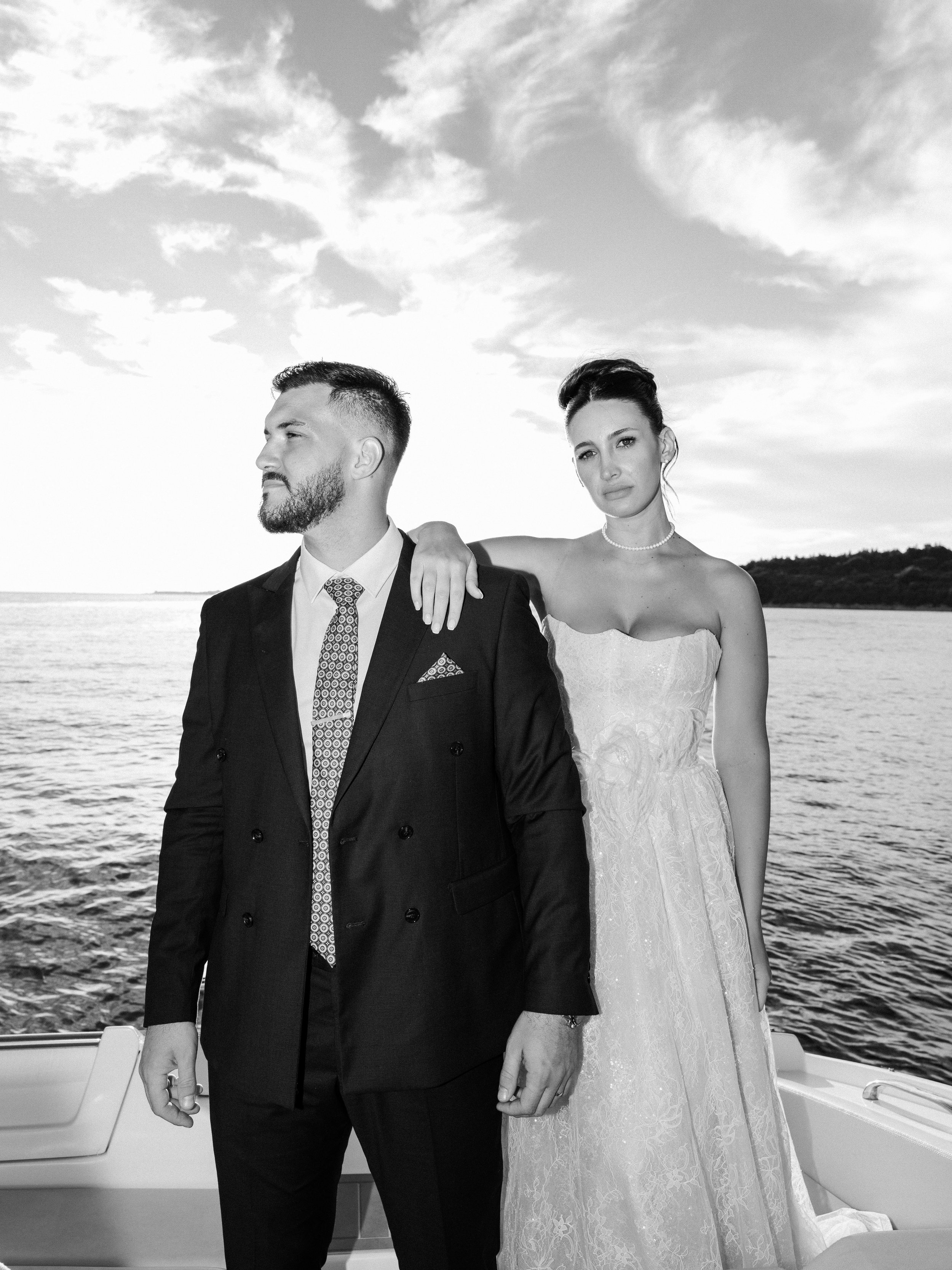 A black and white photo of a couple dressed in wedding attire, standing on a boat by the water with hills and clouds in the background. The man is wearing a suit and tie, and the woman is wearing a strapless wedding dress and pearls, with one arm ove