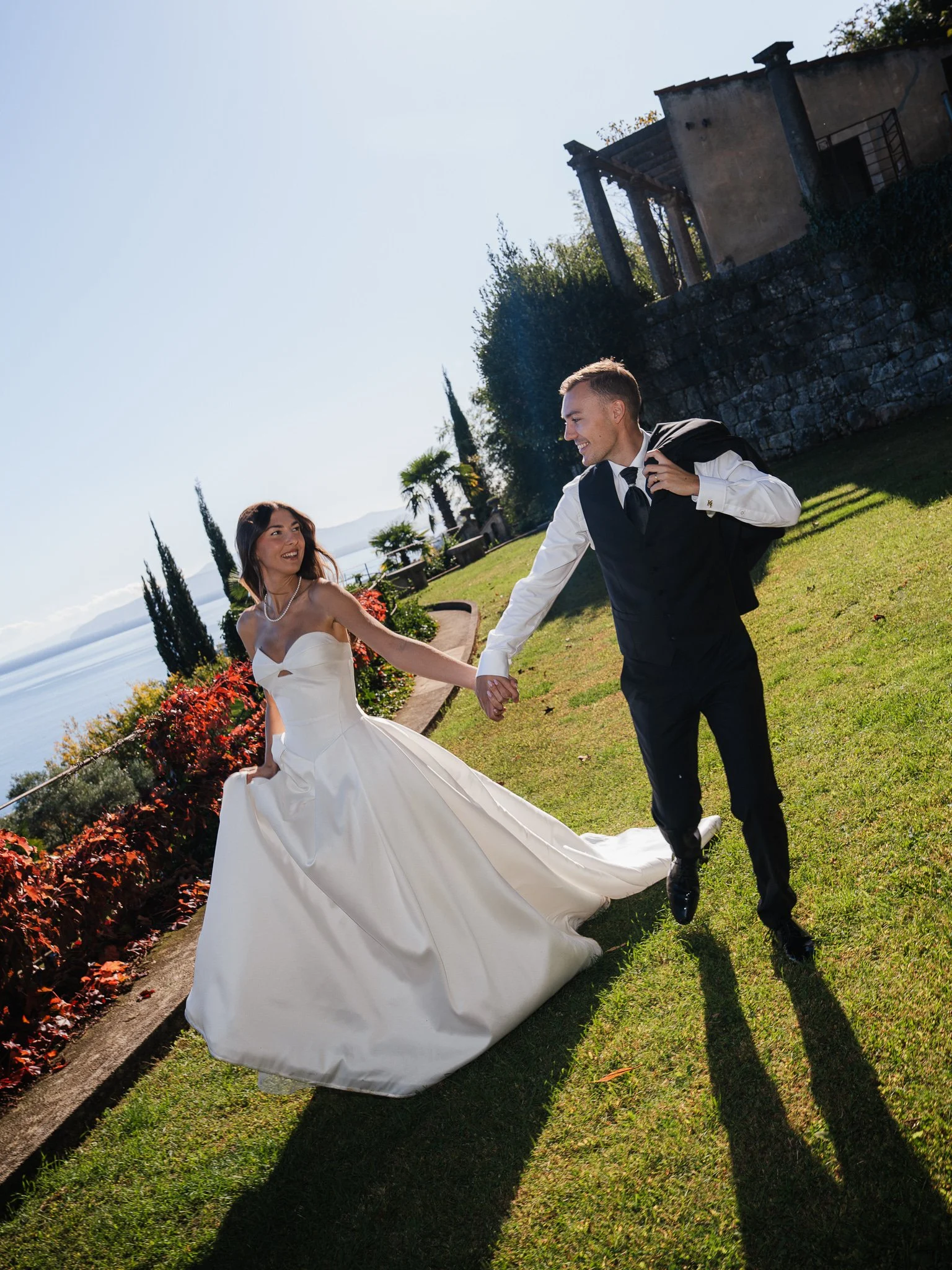 A newlywed couple holding hands and smiling outdoors on a sunny day, with trees, flowers, and a stone building in the background.
