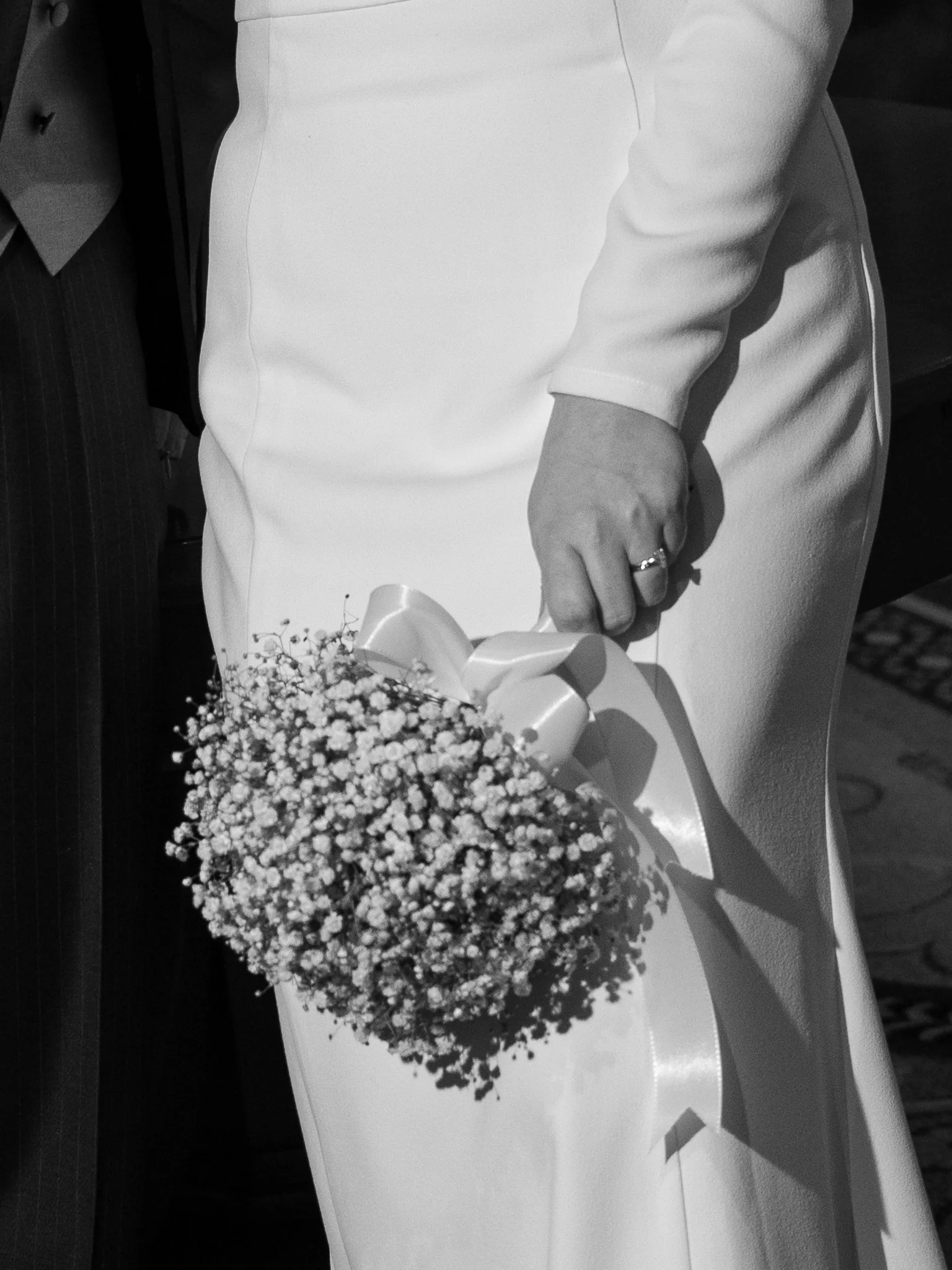 A woman in a white dress holding a bouquet of small white flowers with a ribbon, standing next to a man in dark pinstriped suit.