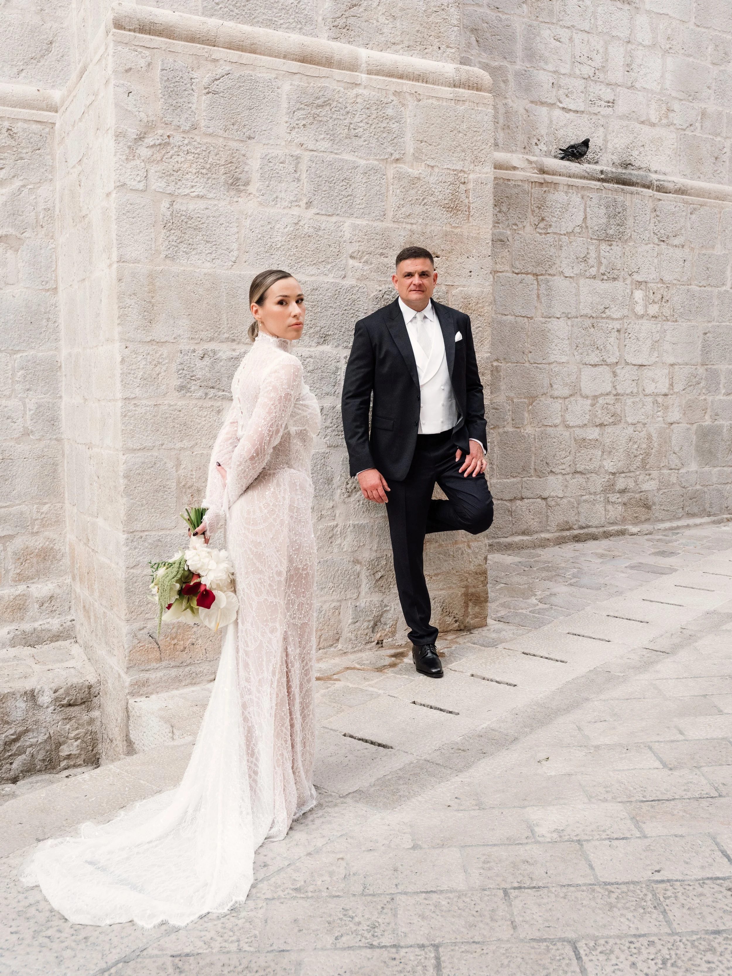 A bride and groom standing against a stone wall outdoors; the bride is holding a bouquet and wearing a lace wedding dress, while the groom is in a black tuxedo with a white shirt and tie, with one leg crossed and hand in pocket.