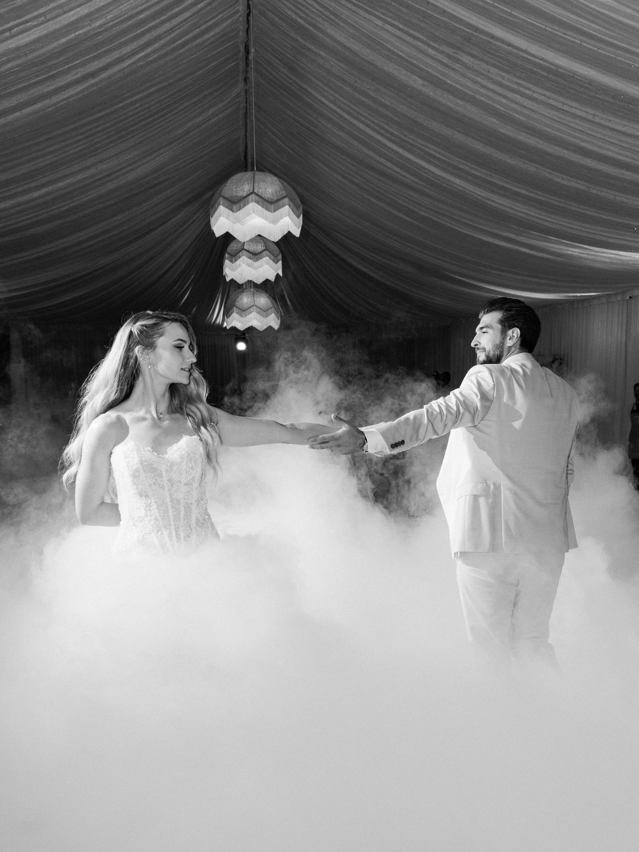 A bride and groom dancing at their wedding reception in a decorated venue with draped fabric ceiling and hanging lamps, surrounded by fog or smoke.