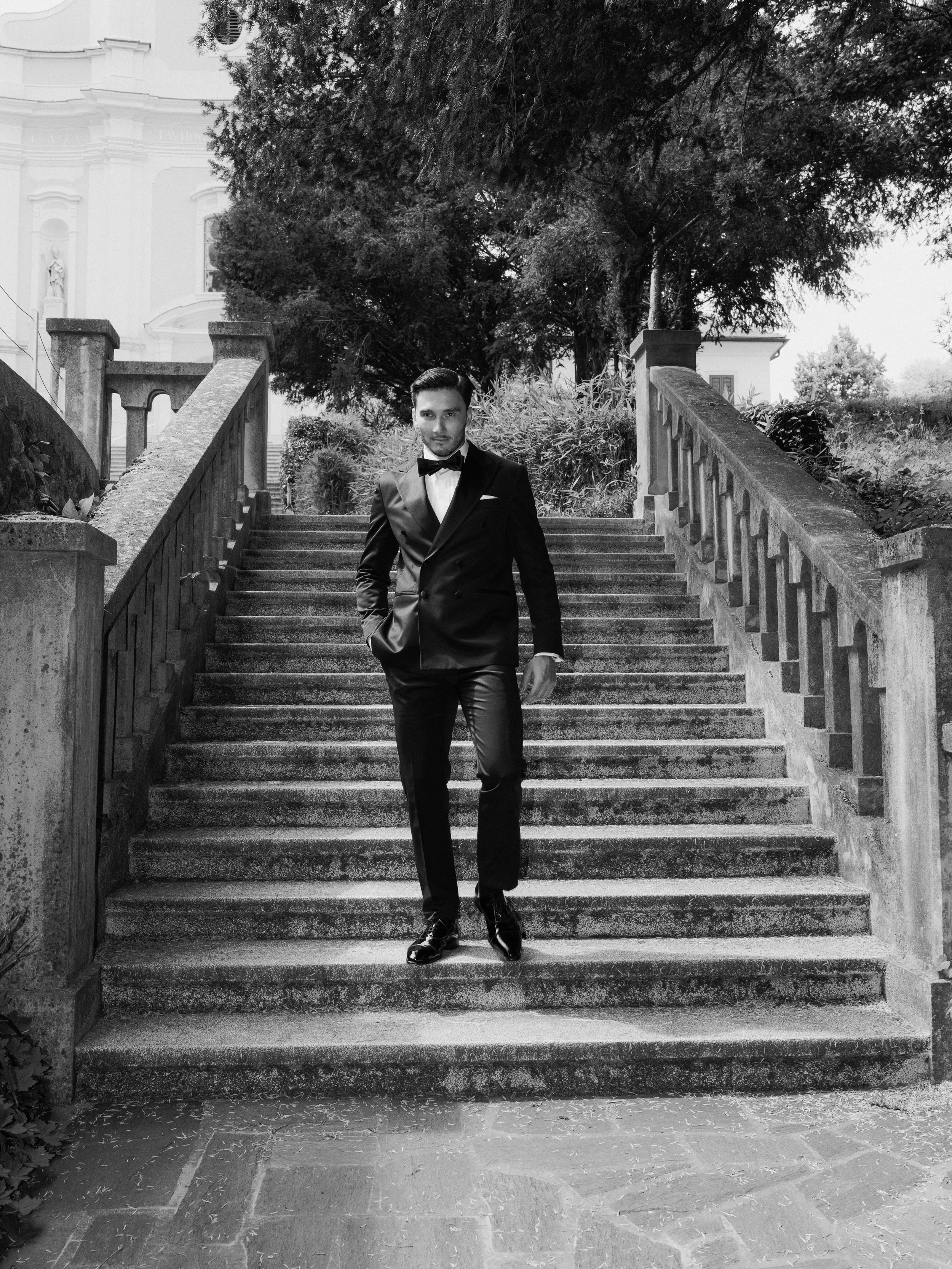 A man in a tuxedo stands on outdoor stairs in front of trees and a building, looking at the camera.