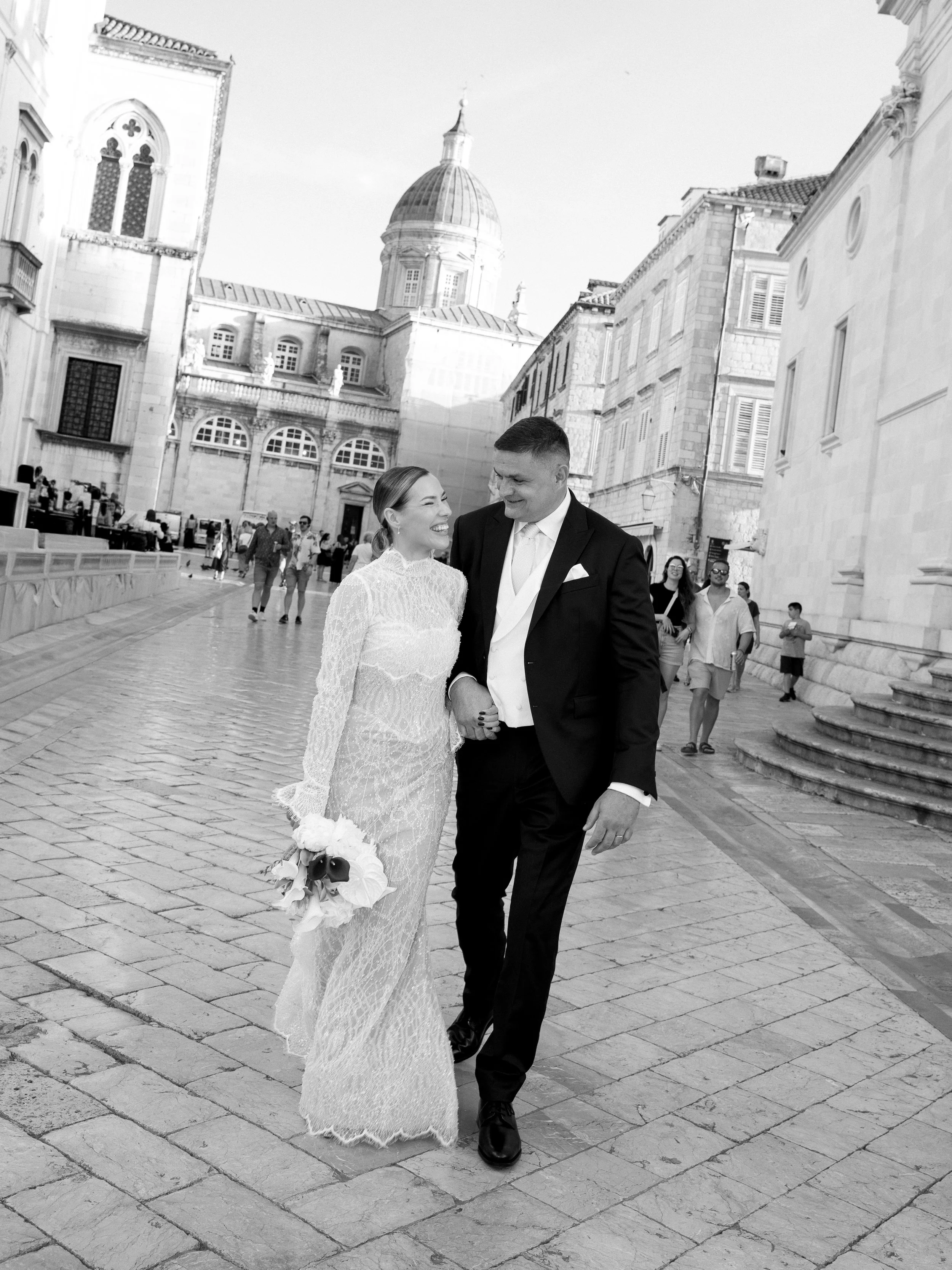 A bride and groom walking hand in hand on a cobblestone street in front of historic European-style buildings, with the bride holding a bouquet of flowers, and the groom dressed in a tuxedo, smiling at each other.