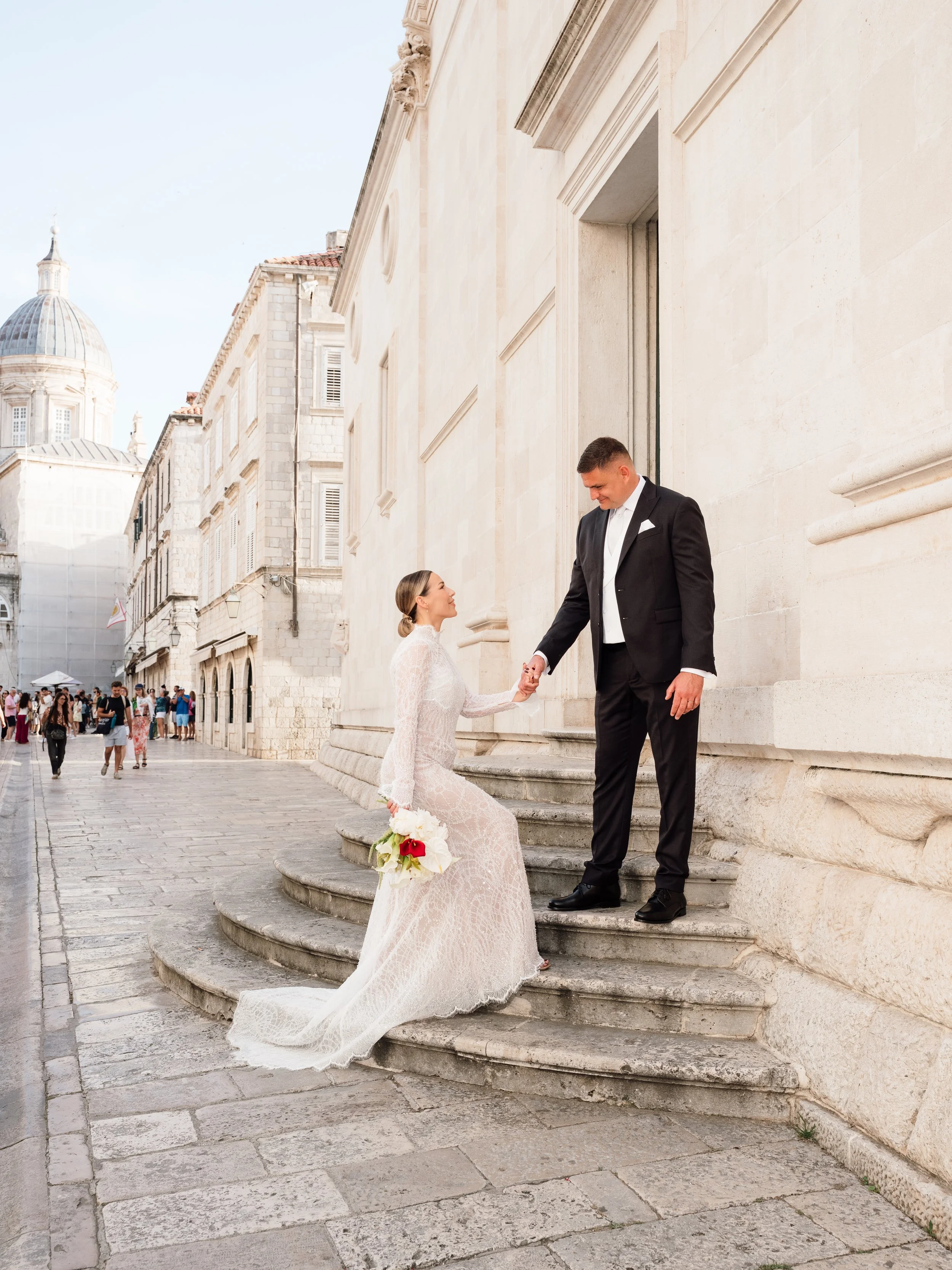 A bride and groom are standing on stone steps outside a historic building. The bride is kneeling and holding a bouquet of white and red flowers, while the groom is standing above her, gently holding her hand. The bride is wearing a long, white lace w
