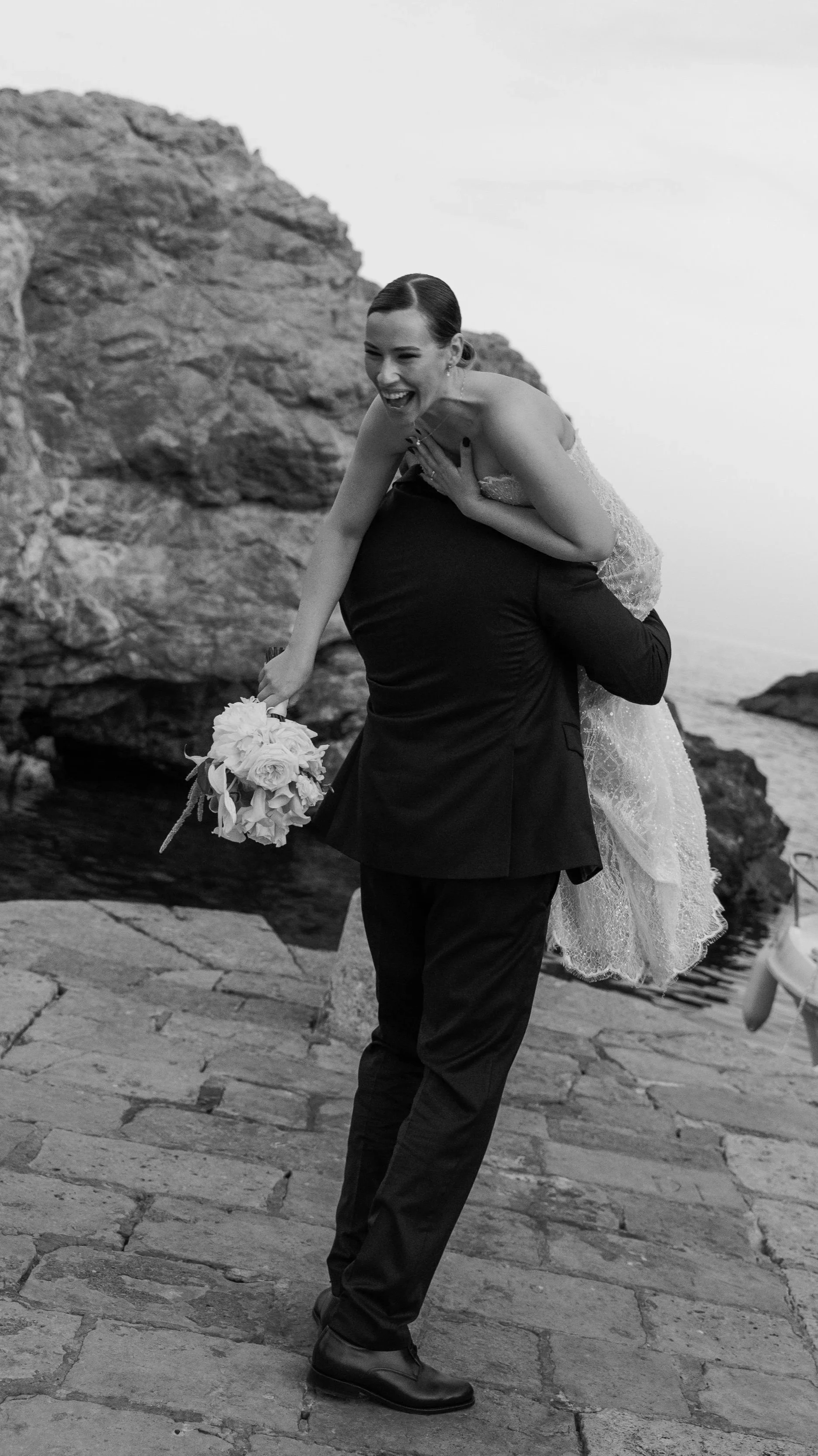 A bride and groom are celebrating on a rocky outdoor location, with the bride laughing and holding a bouquet of flowers. The groom holds the bride over his shoulder.