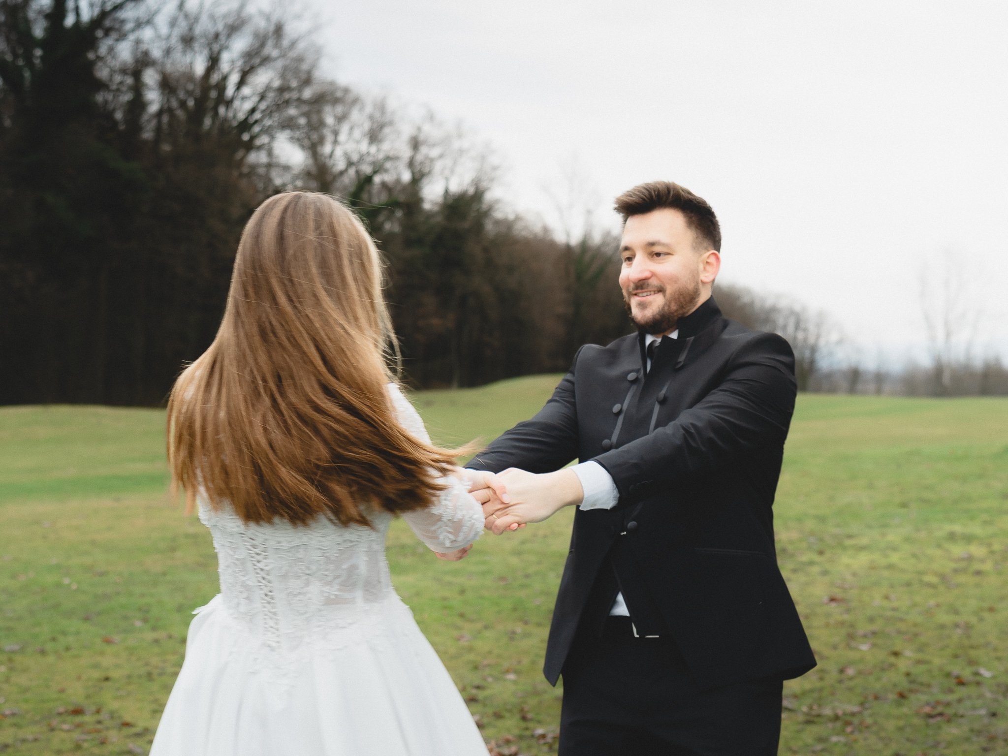 A couple is holding hands and smiling outdoors on a grassy field with trees in the background. The woman is wearing a white dress and has long, brown hair; the man is wearing a black tuxedo and has short brown hair and a beard.