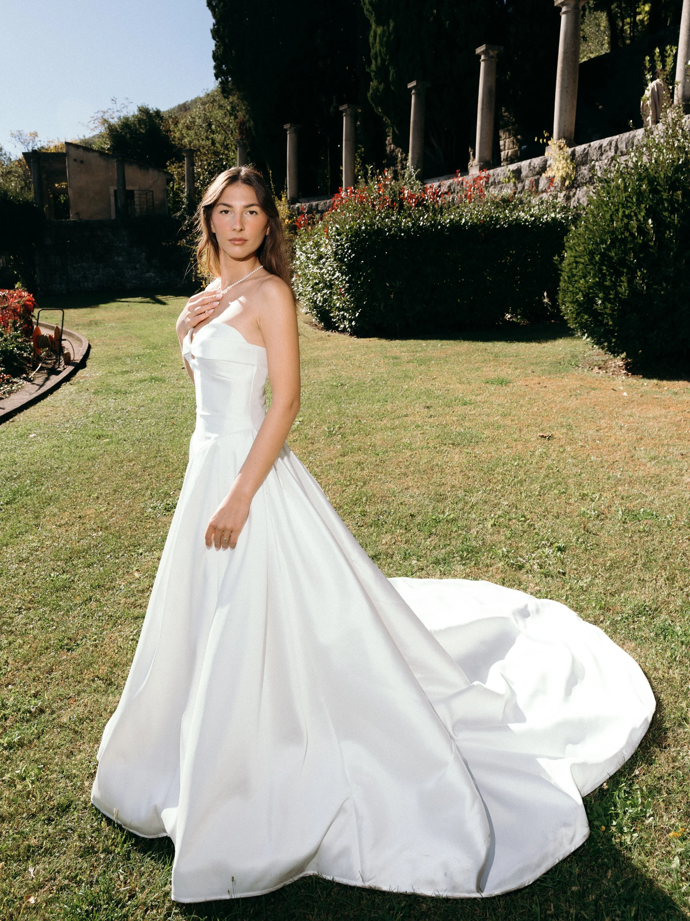 A woman in a white wedding dress standing outdoors on a grassy lawn with greenery and stone columns in the background.