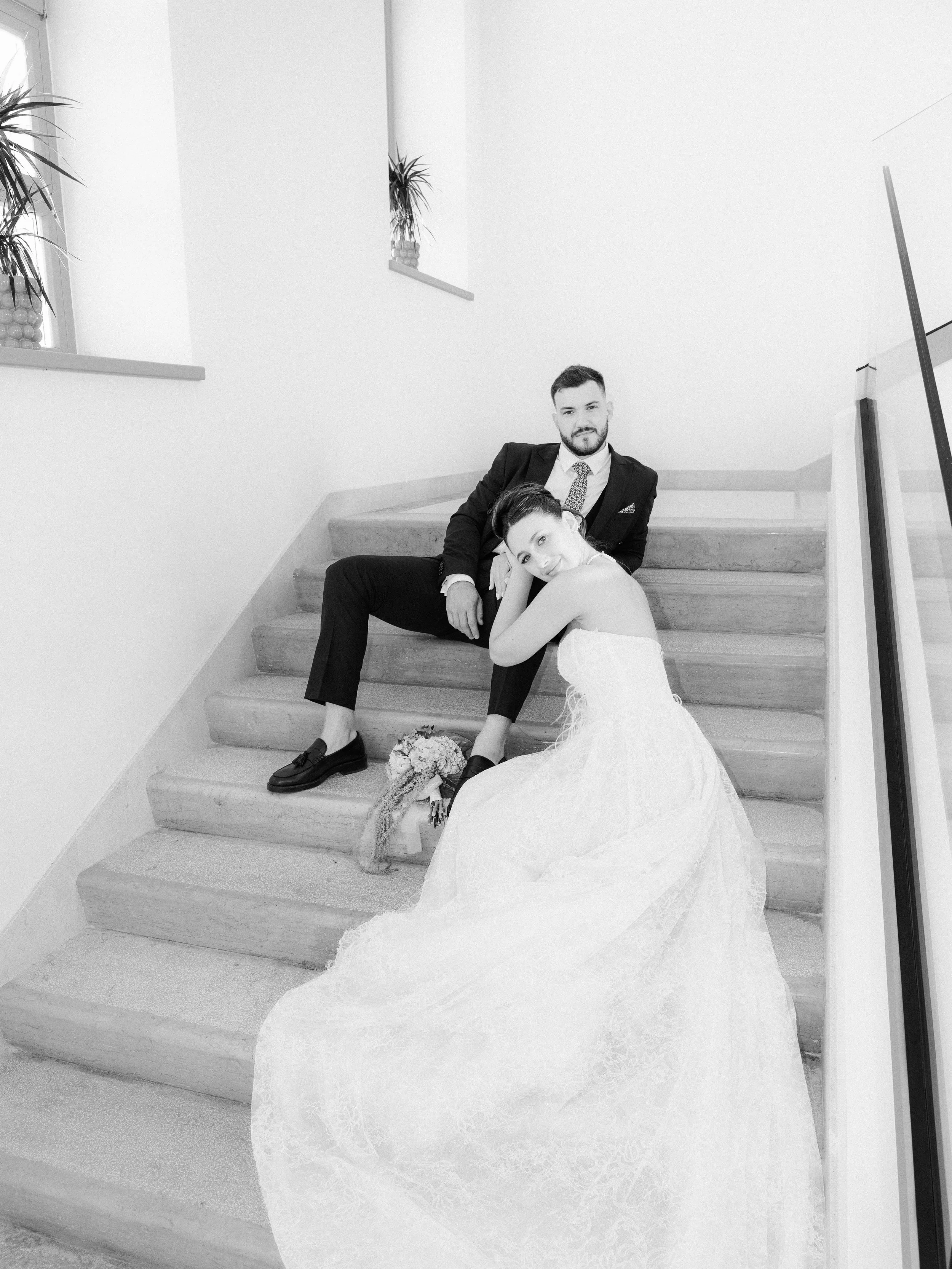 A man in a suit and a woman in a wedding dress sitting on stairs in a minimalist interior, with a bouquet of flowers on the floor.