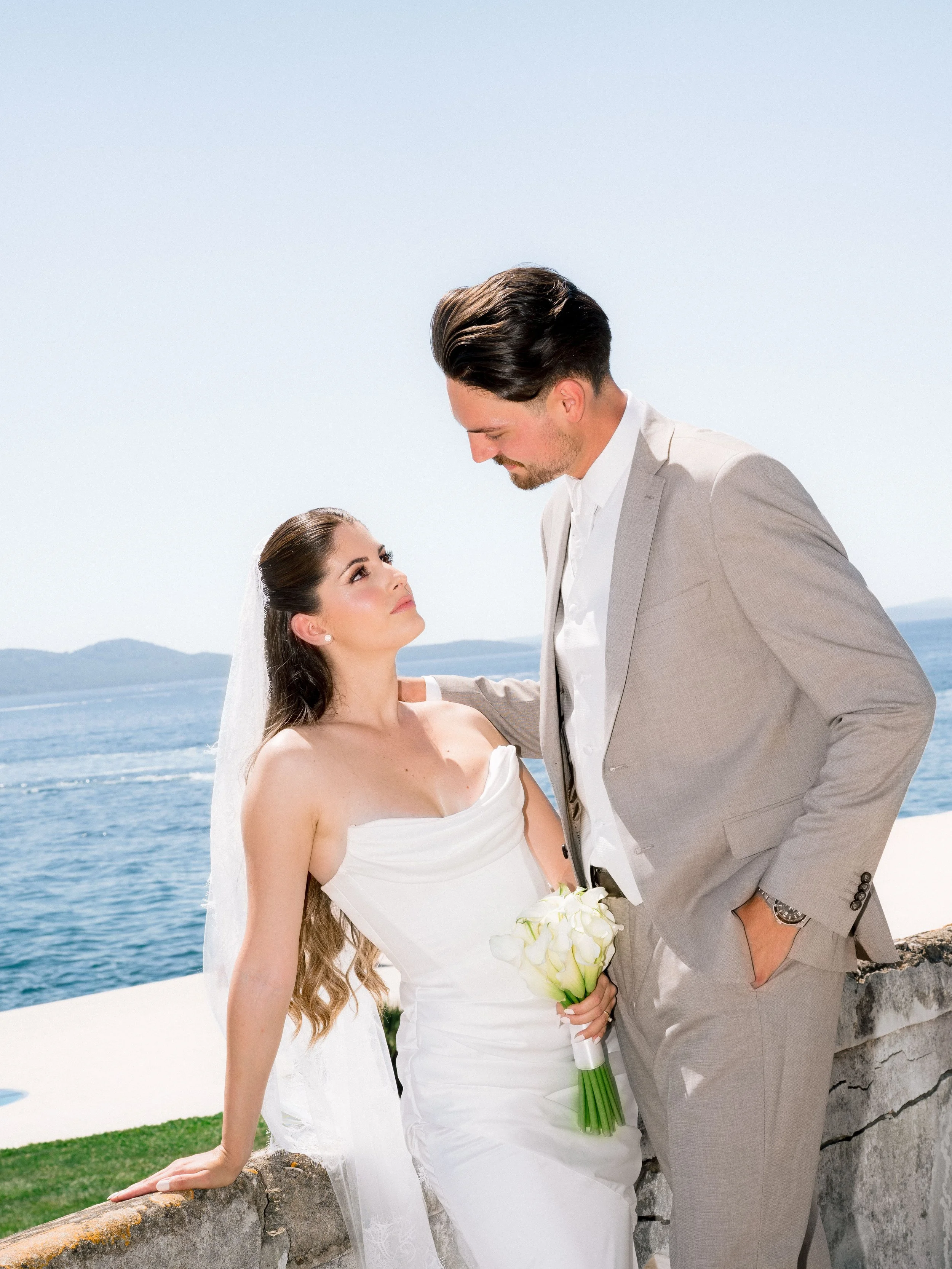 A bride and groom standing near a rocky waterfront, with the bride holding a bouquet of white flowers and looking up at the groom, who is looking down at her. The background shows a body of water and distant mountains under a clear sky.