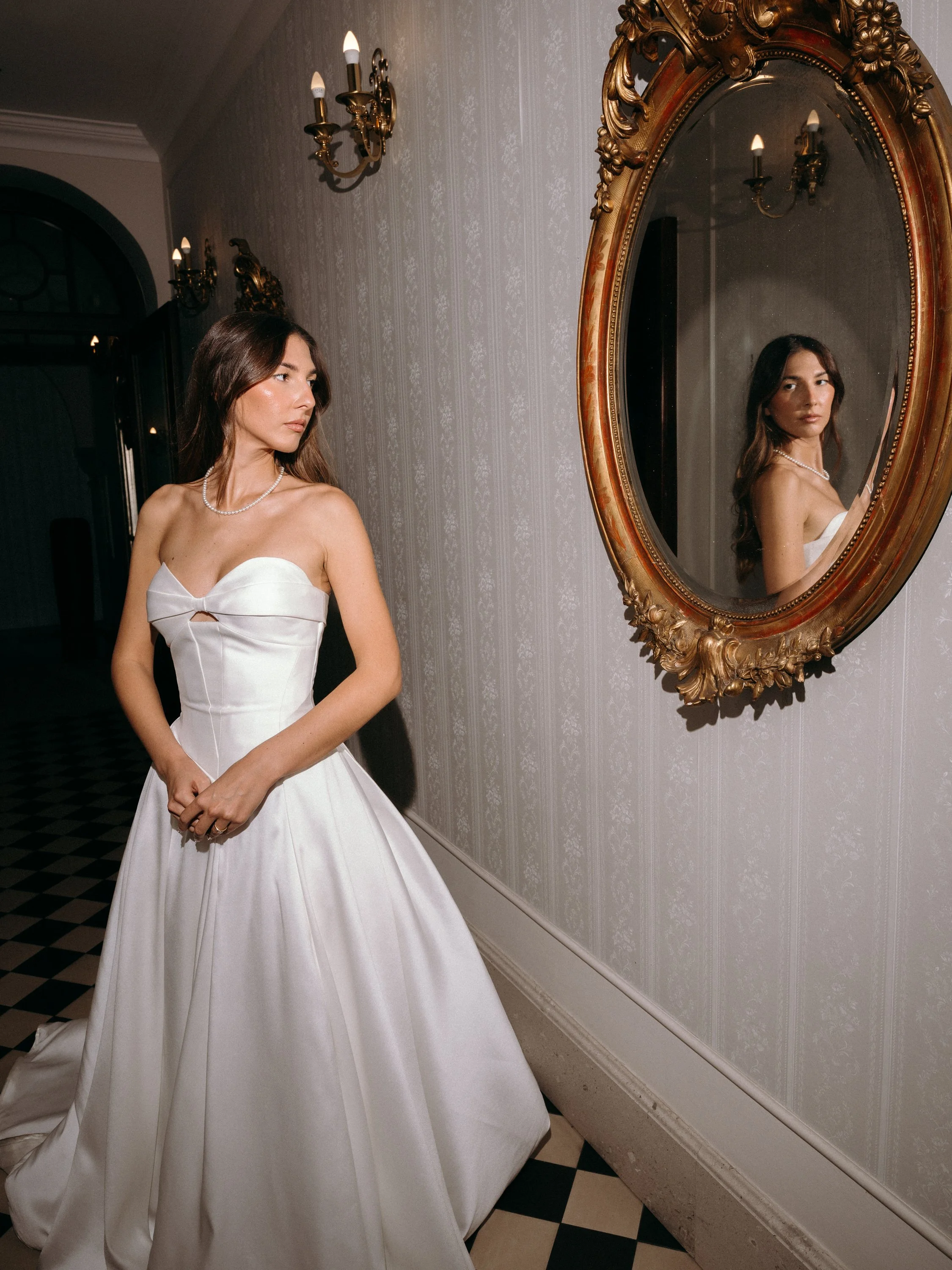 A woman in an elegant white strapless dress and pearl necklace stands in front of an ornate gold-framed mirror on a patterned black-and-white checkered floor. The mirror reflects her face and upper body.