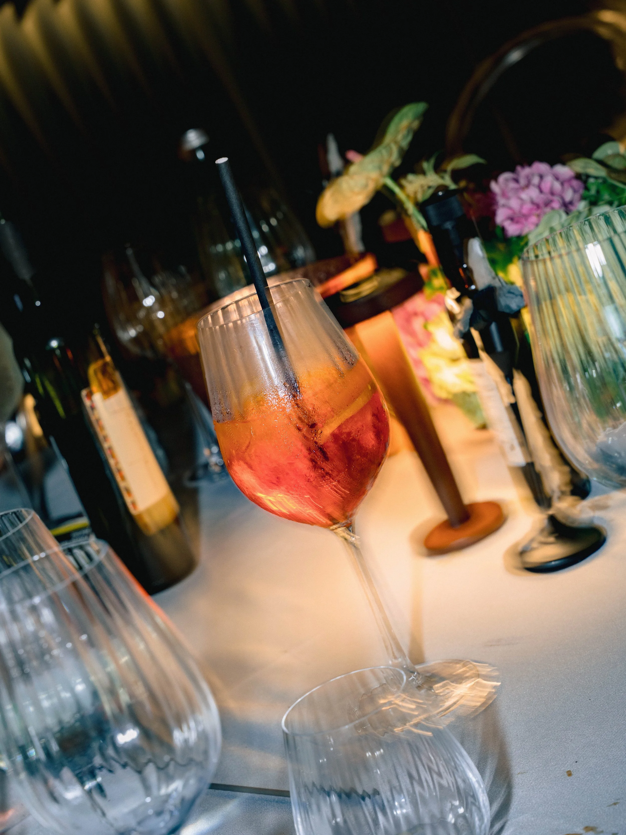 Close-up of two glasses with colorful drinks and black straws on a table, with blurred background including bottles, flowers, and tableware.