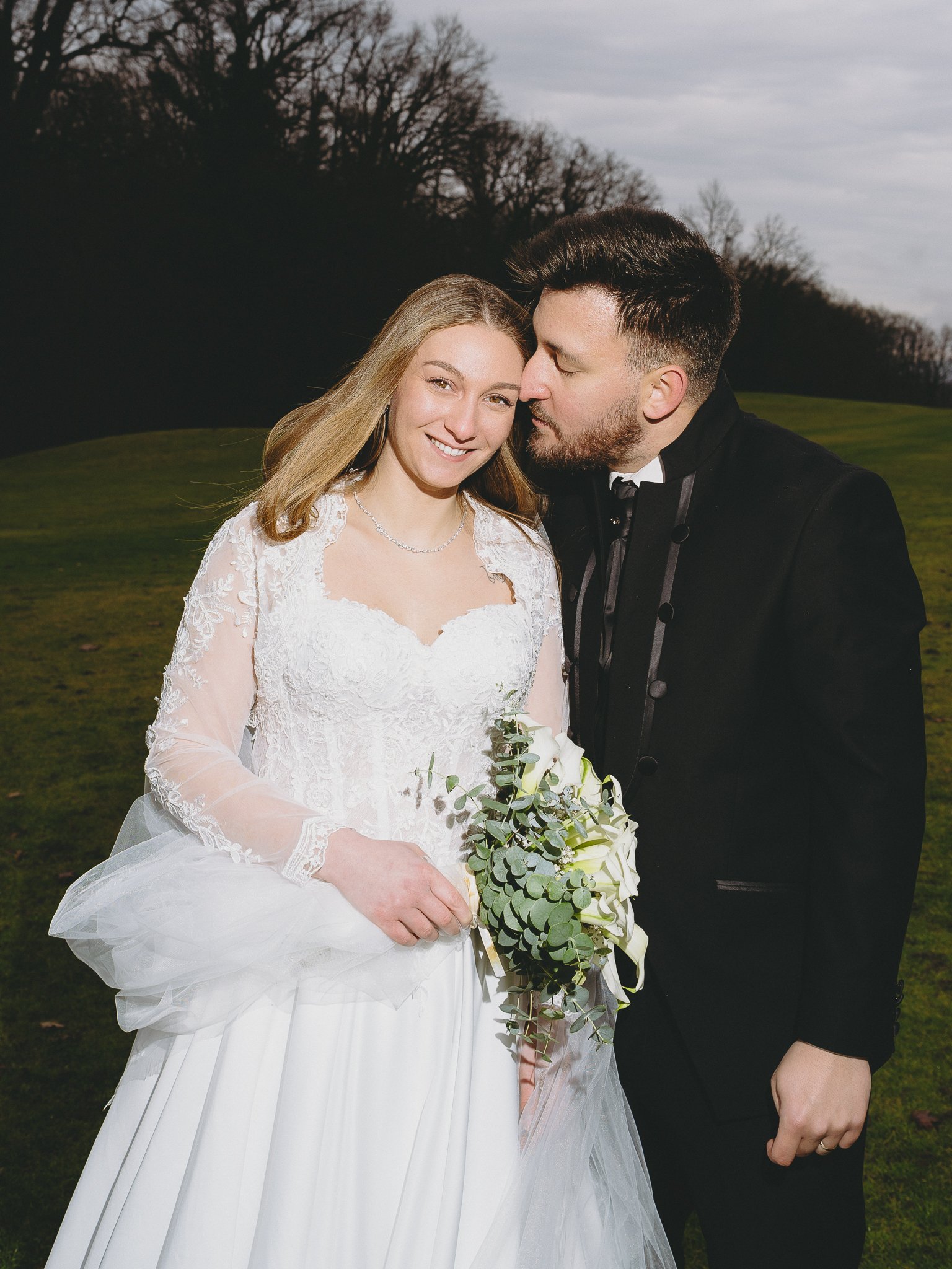 A bride and groom standing outdoors on a cloudy day, with the groom leaning in close to the bride. The bride is holding a bouquet of white flowers and greenery, and is dressed in a white lace wedding gown with long sleeves. The groom is wearing a bla