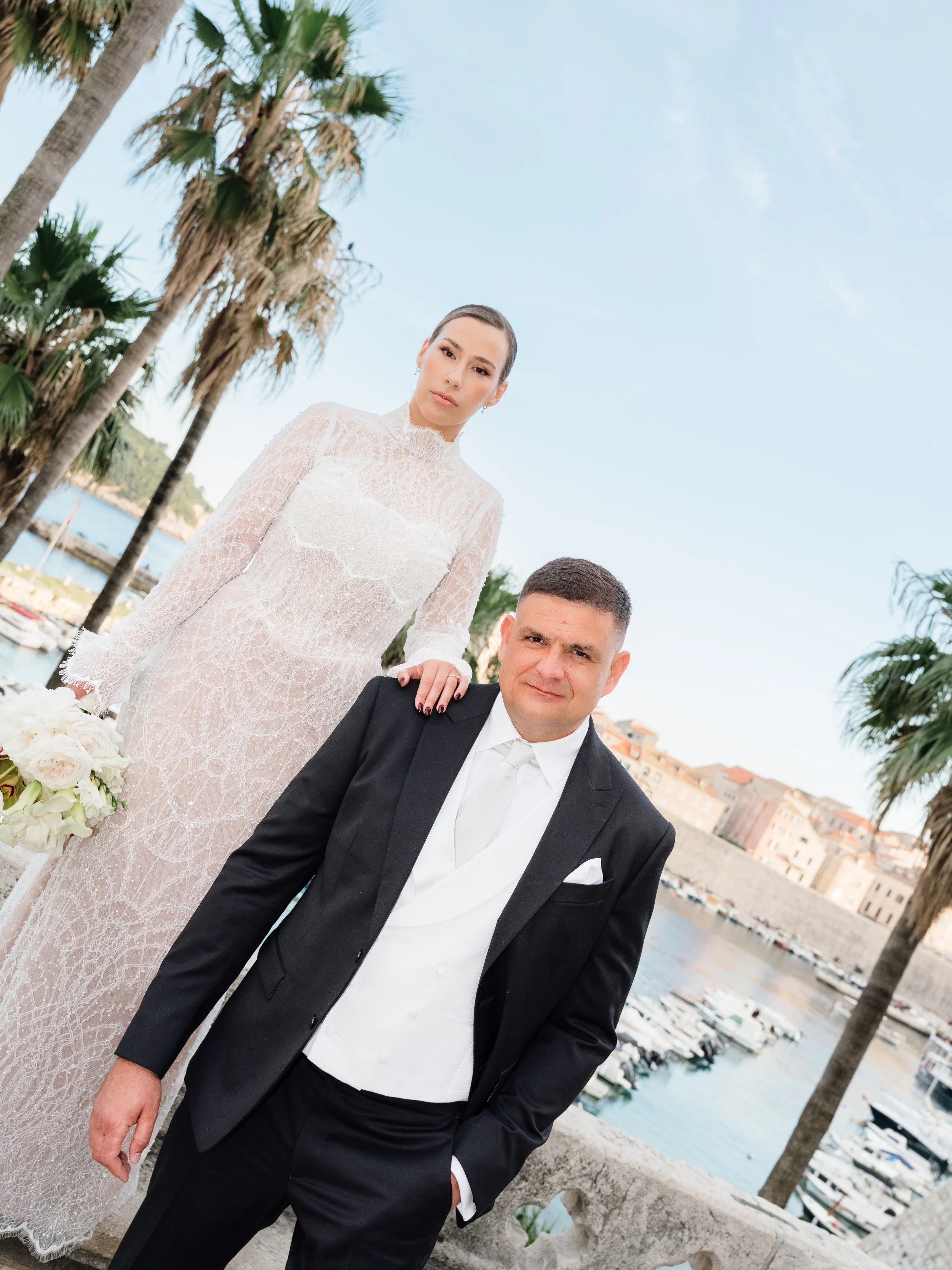 A bride in a white lace wedding dress stands beside a groom in a black tuxedo with a white vest and tie, outdoors near water and palm trees, with a cityscape background.