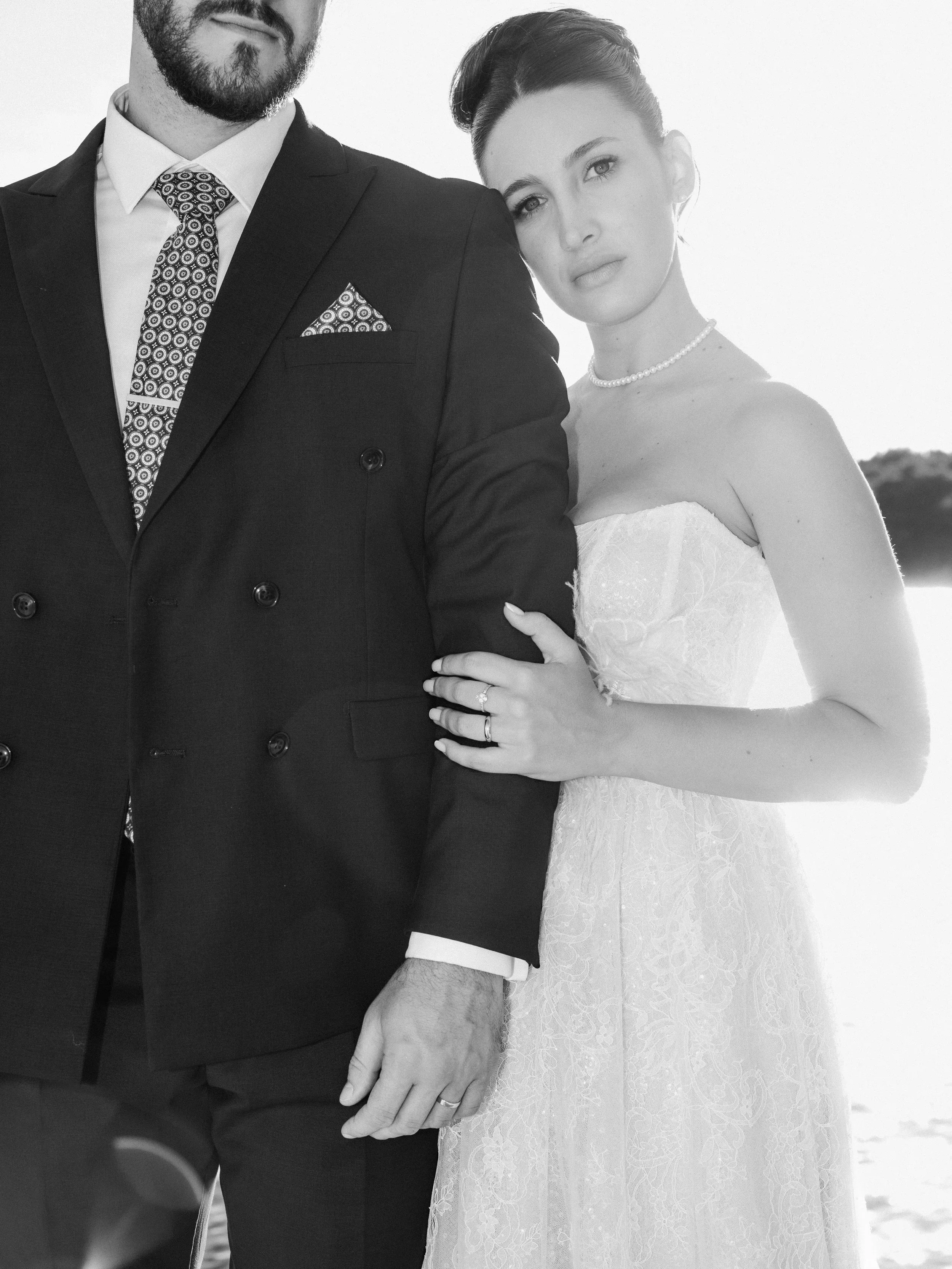 A black and white photo of a couple on their wedding day, the woman wearing a lace wedding dress, pearl necklace, and wedding ring, with her hand resting on the man's chest. The man is dressed in a dark suit with a patterned tie and pocket square, an