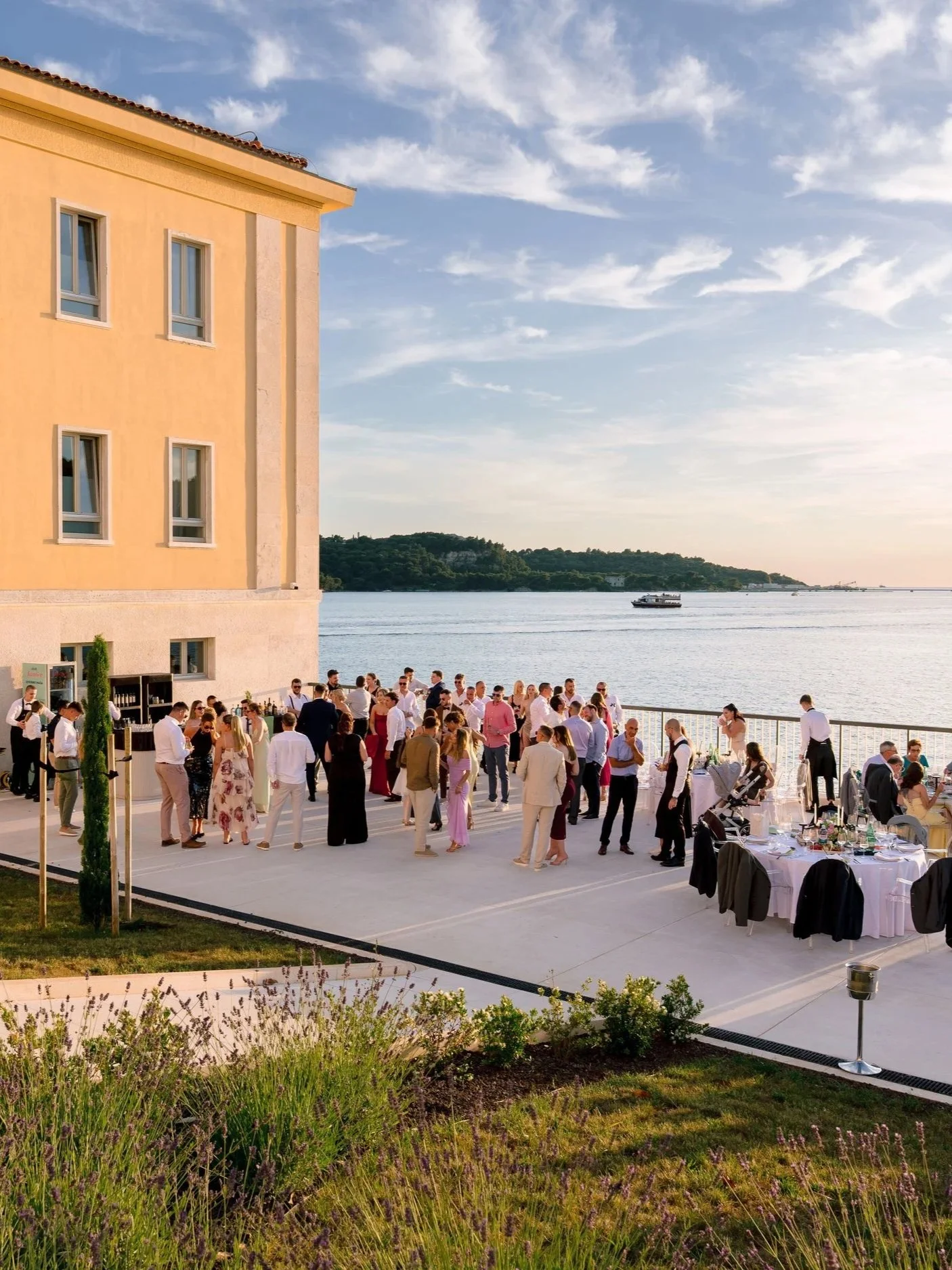 A large gathering of people dressed formally is hosting an outdoor event by the water, with tables set up and a yellow building with windows on the left side of the image. The sky is partly cloudy with a boat on the water in the background.