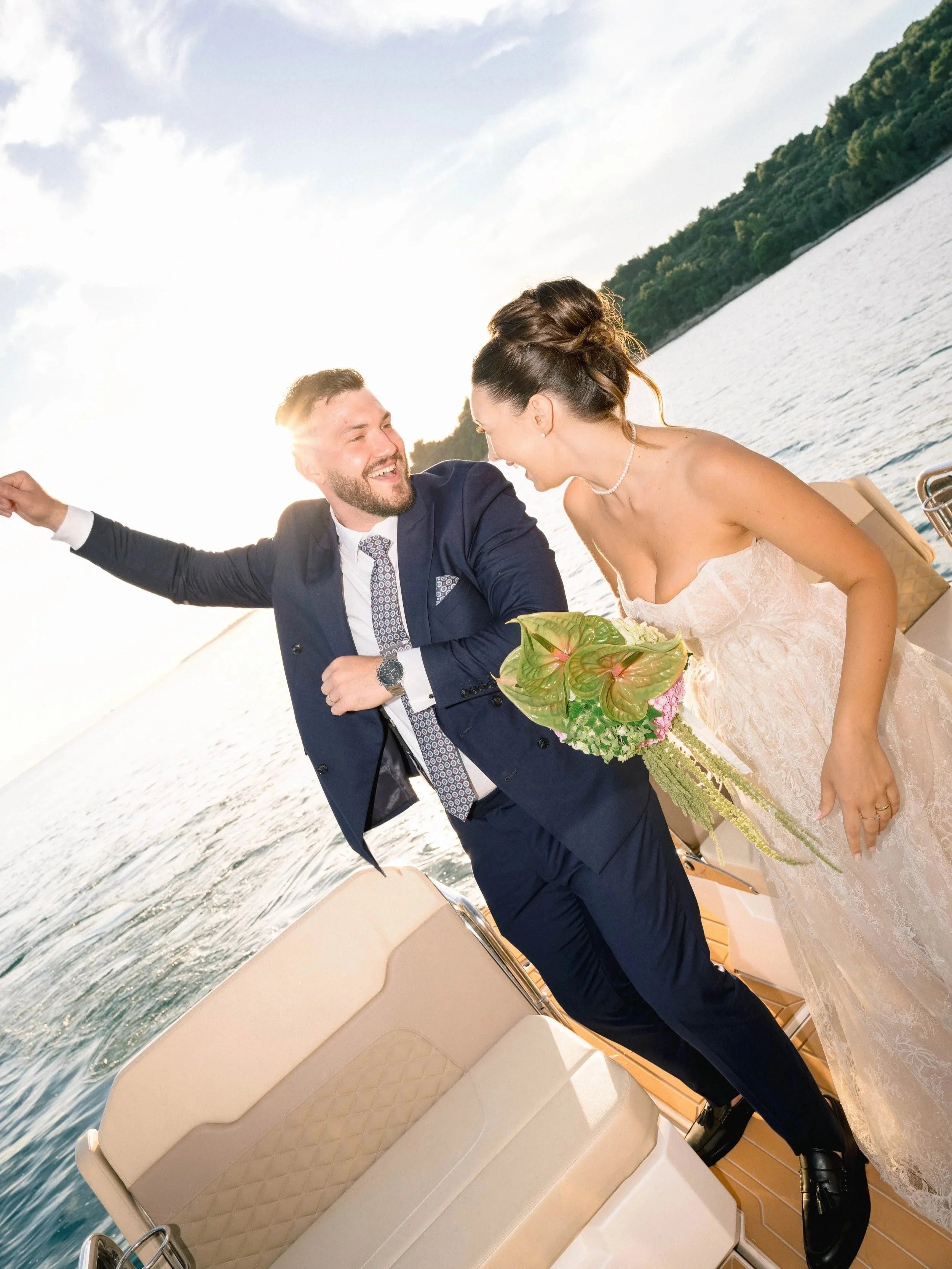 A bride and groom on a boat at sunset, smiling and laughing together as they look at each other, with water and green trees in the background.