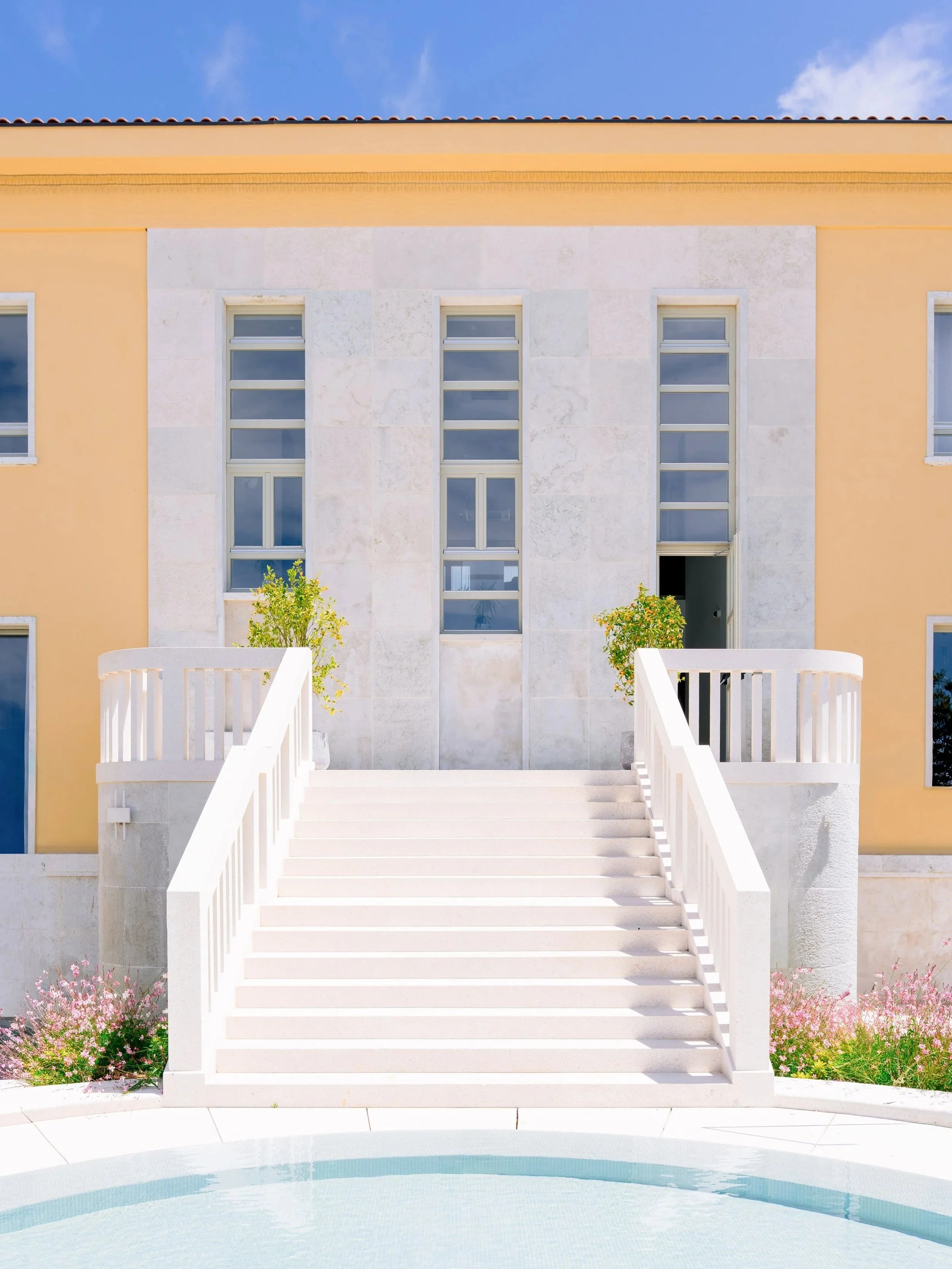 Exterior view of a modern house with white stairs leading to the entrance, yellow walls, multiple windows, and a swimming pool in the foreground, with clear blue sky and pink flowers at the base.