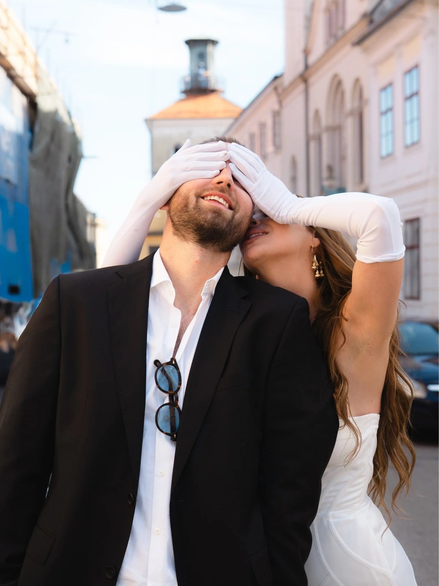 A man in a black suit with sunglasses hanging from his shirt standing outdoors as a woman in a white dress playfully covers his eyes with white gloves, with buildings and a street in the background.