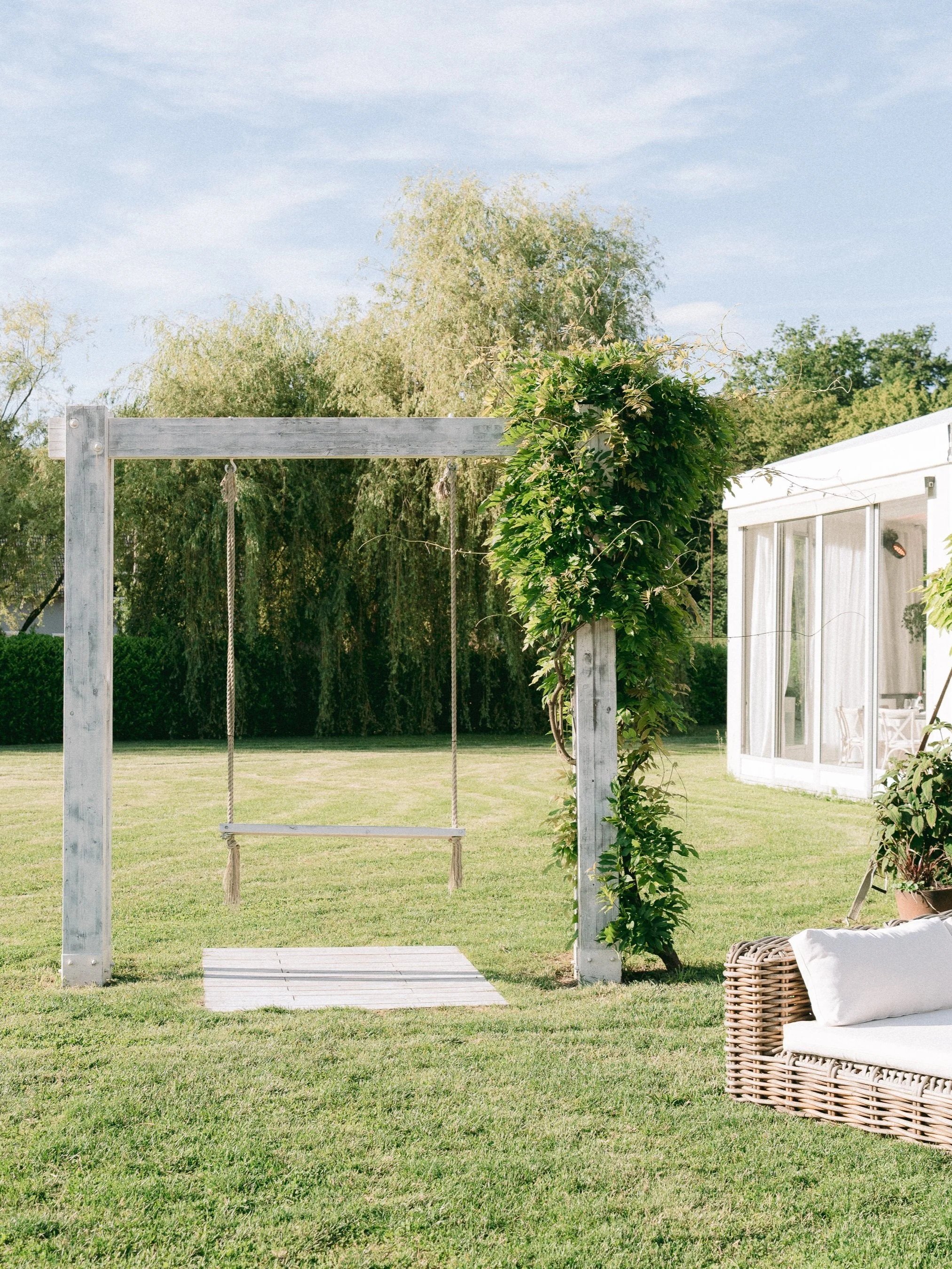 Outdoor backyard with a wooden swing set, greenery, a modern white house with large windows, and outdoor furniture.