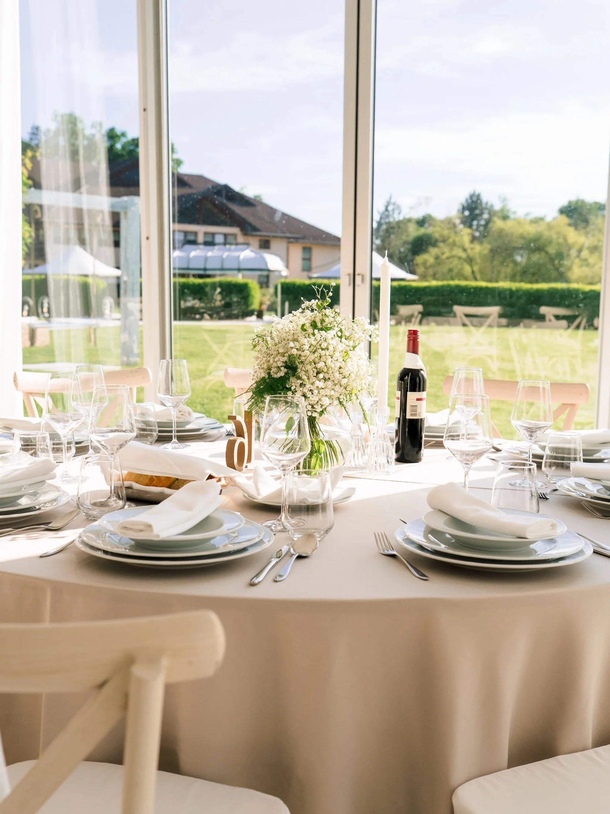 A round dining table set for a formal meal indoors near large windows with a view of a green garden and a house outside. The table has a beige tablecloth, white plates, glasses, silverware, napkins, and a floral centerpiece with white flowers and a c